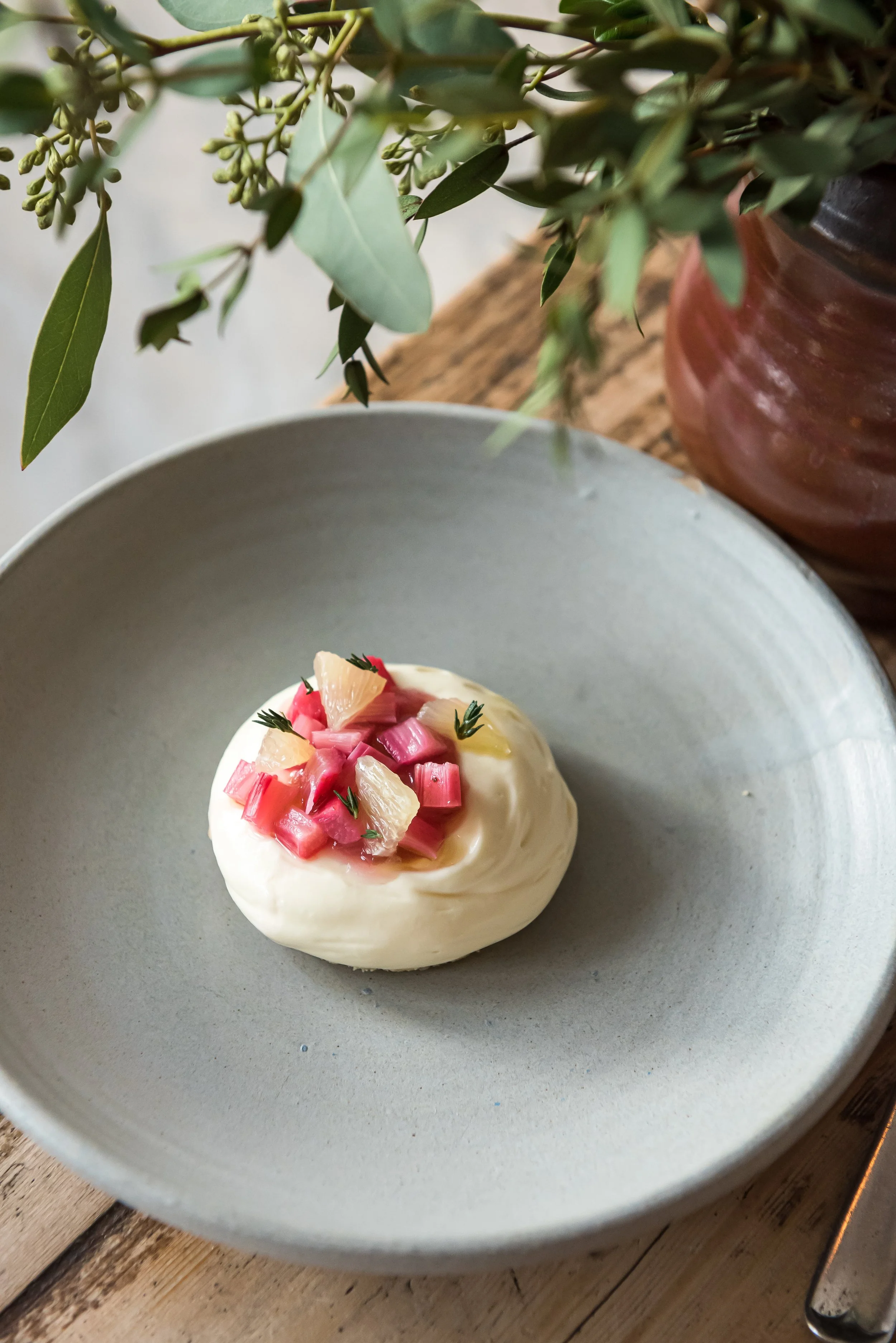 A bowl with a soft, creamy dessert garnished with chopped pink and yellow fruit and herbs, placed on a wooden surface with a potted plant nearby.