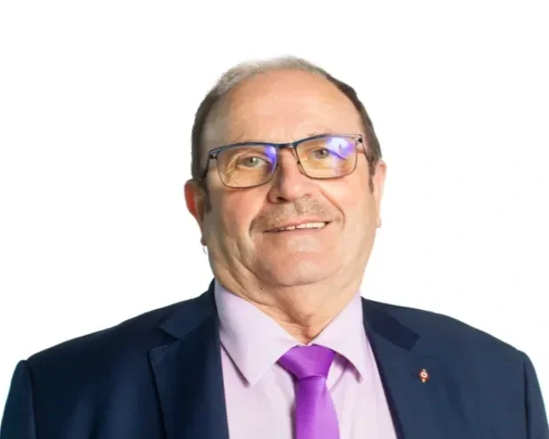 Un homme portant un costume foncé, une chemise rose pâle, une cravate violette, et des lunettes, souriant devant un fond blanc.