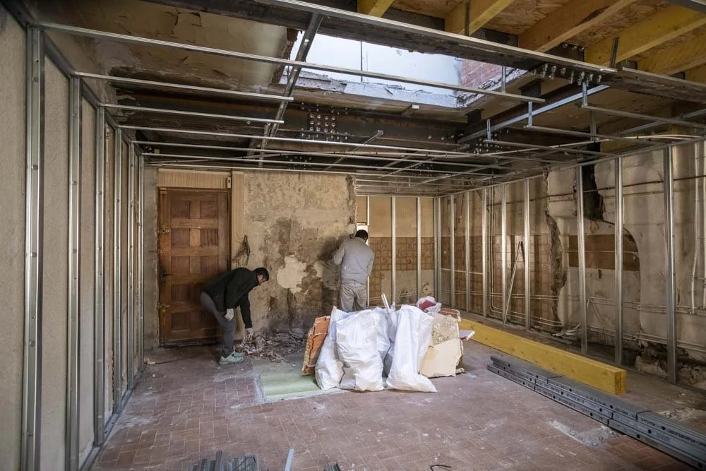 Deux ouvriers travaillent dans une pièce en rénovation, avec une structure métallique en place et des murs partiellement démolis.