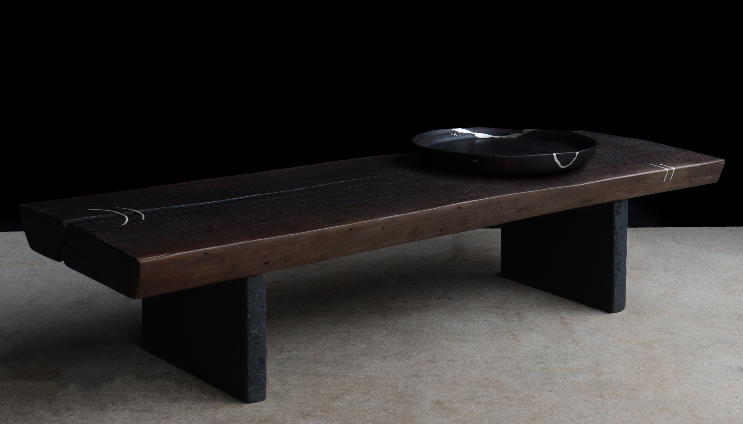 A dark wooden table with black stone supports and a black ceramic plate on top, set against a black background.