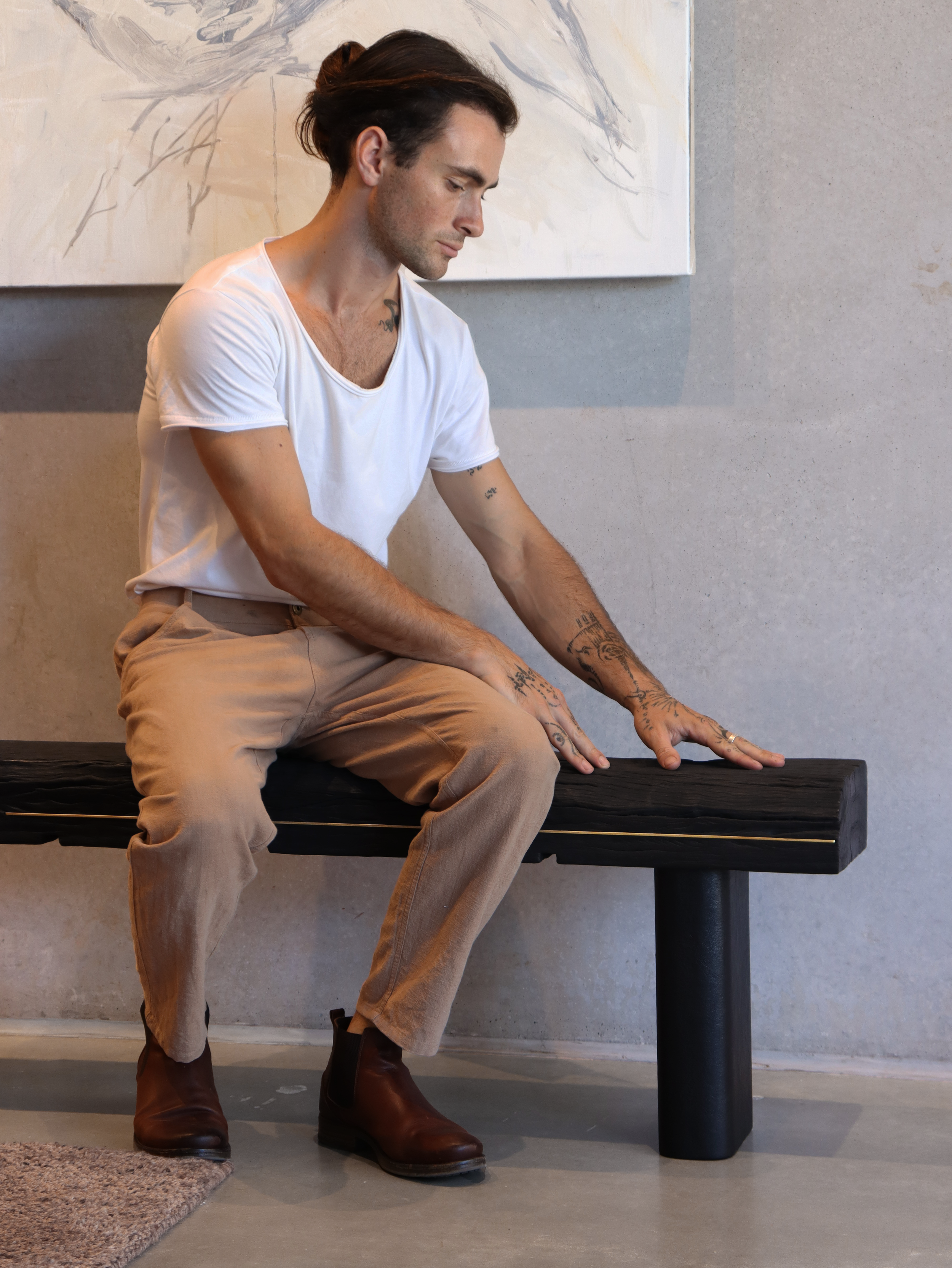A young man with tattoos, wearing a white t-shirt and beige pants, sitting on a black bench with his hands resting on the seat, looking down with a pensive expression, in a room with neutral-colored walls and a large abstract painting.