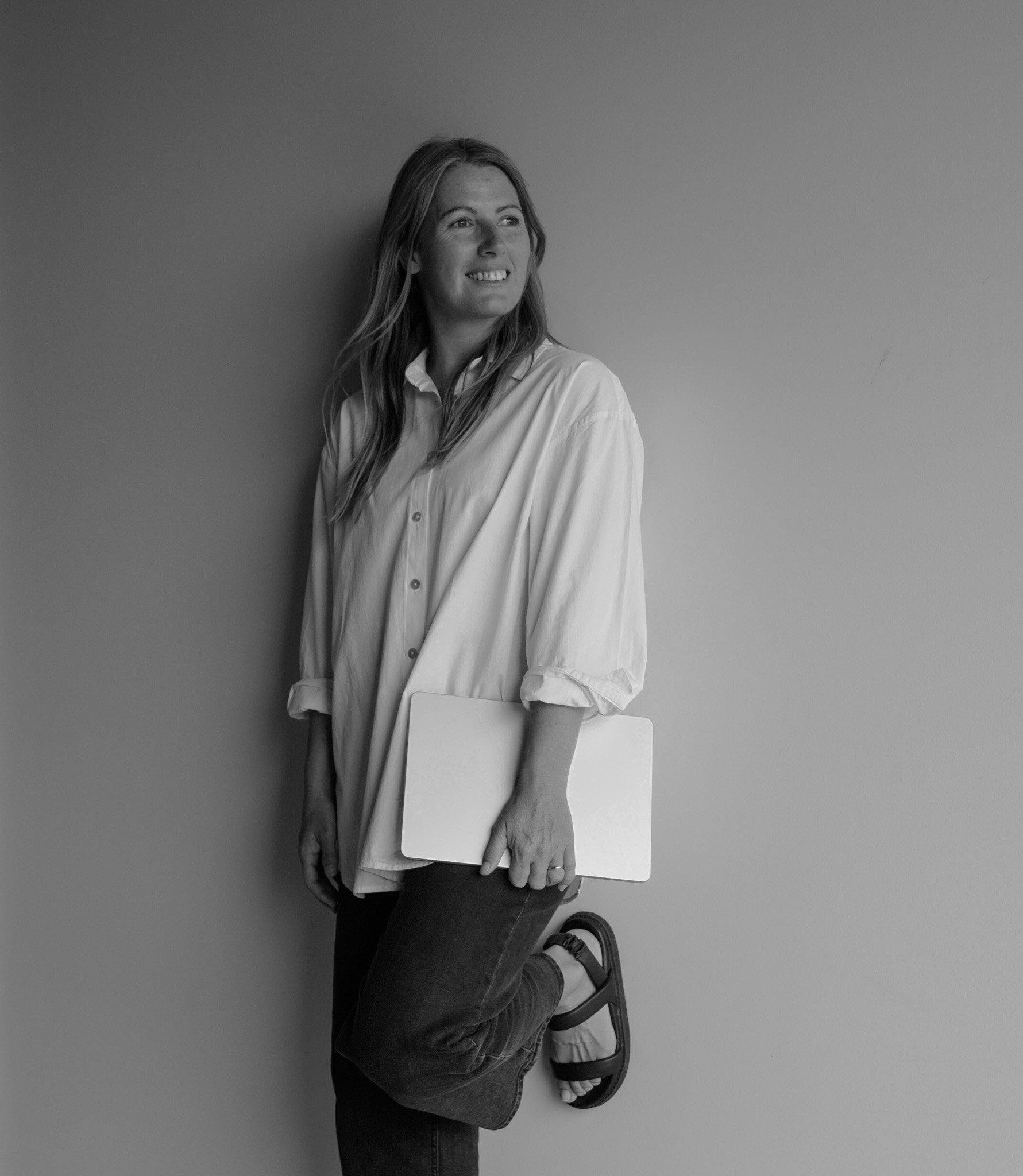Black and white photo of a woman with long hair wearing a light-colored shirt and dark jeans. She is sitting against a plain wall, holding a closed laptop on her lap. She is smiling and looking slightly off to the side.