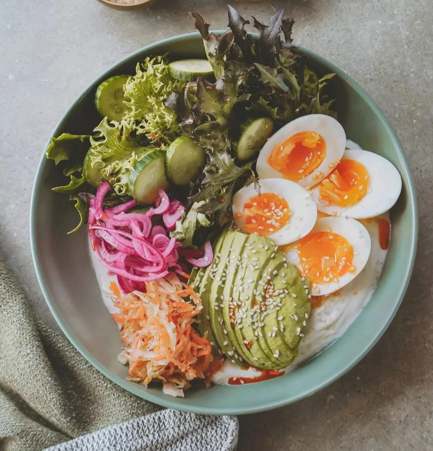 A bowl of salad featuring sliced boiled eggs, shredded carrots and cabbage, sliced cucumbers, leafy greens, and sliced avocado, garnished with sesame seeds.