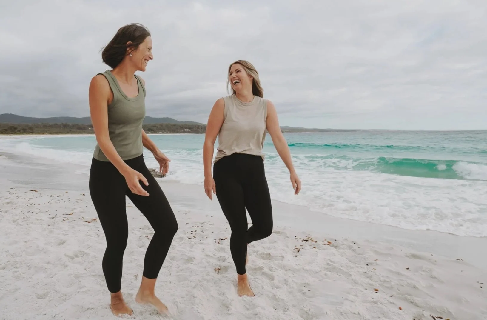 Two women laughing and walking on the beach in casual clothing, with ocean waves and cloudy sky in the background.