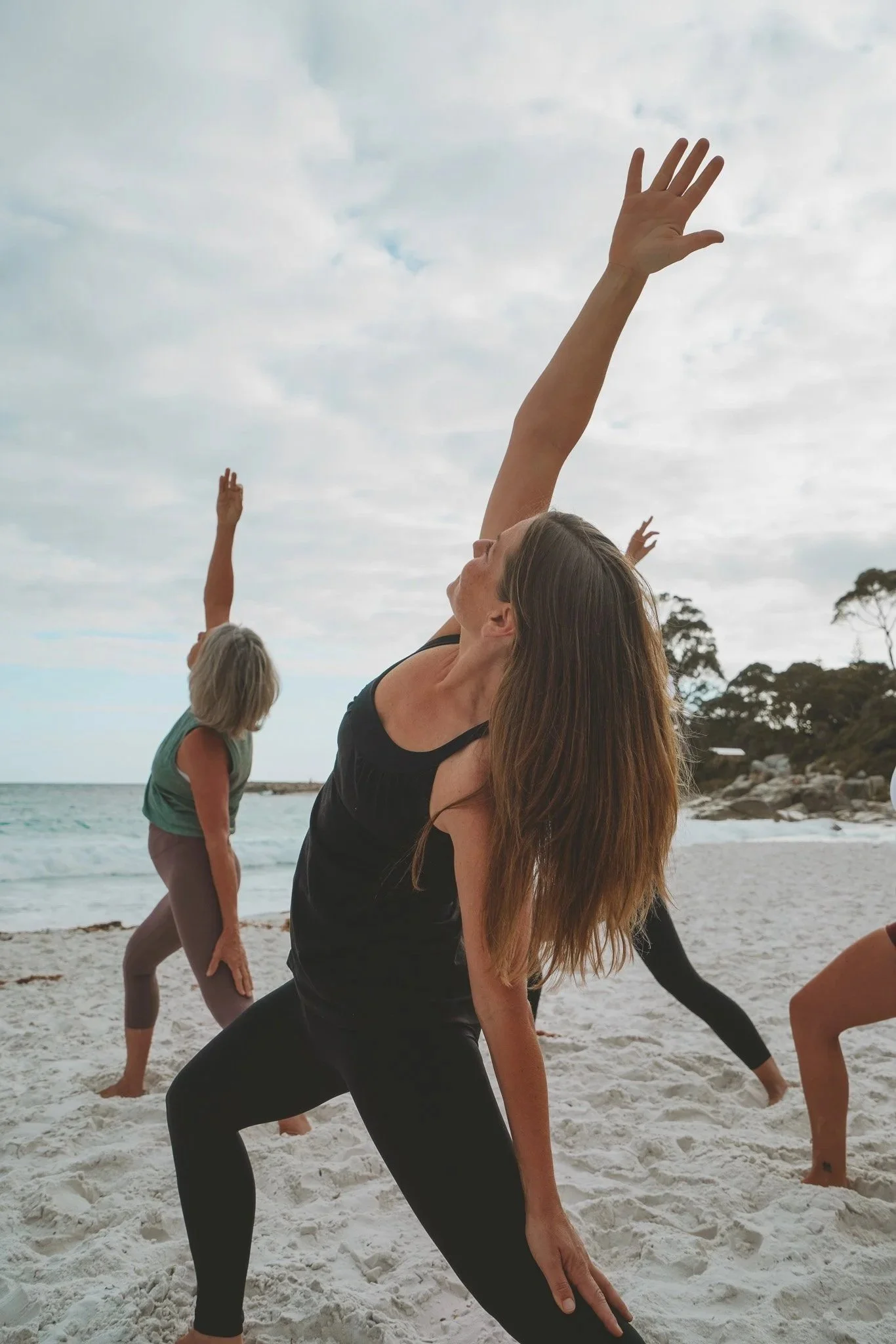 Four women practicing yoga outdoors, raising their arms towards the sky in a circle, looking up, under a cloudy sky with trees in the background.