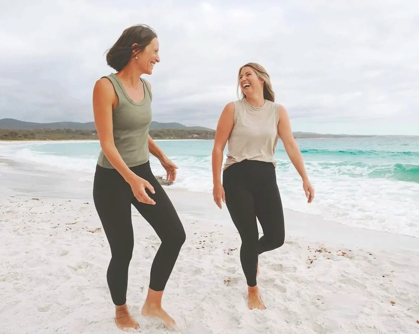 Two women walking and laughing on a sandy beach with ocean waves in the background.
