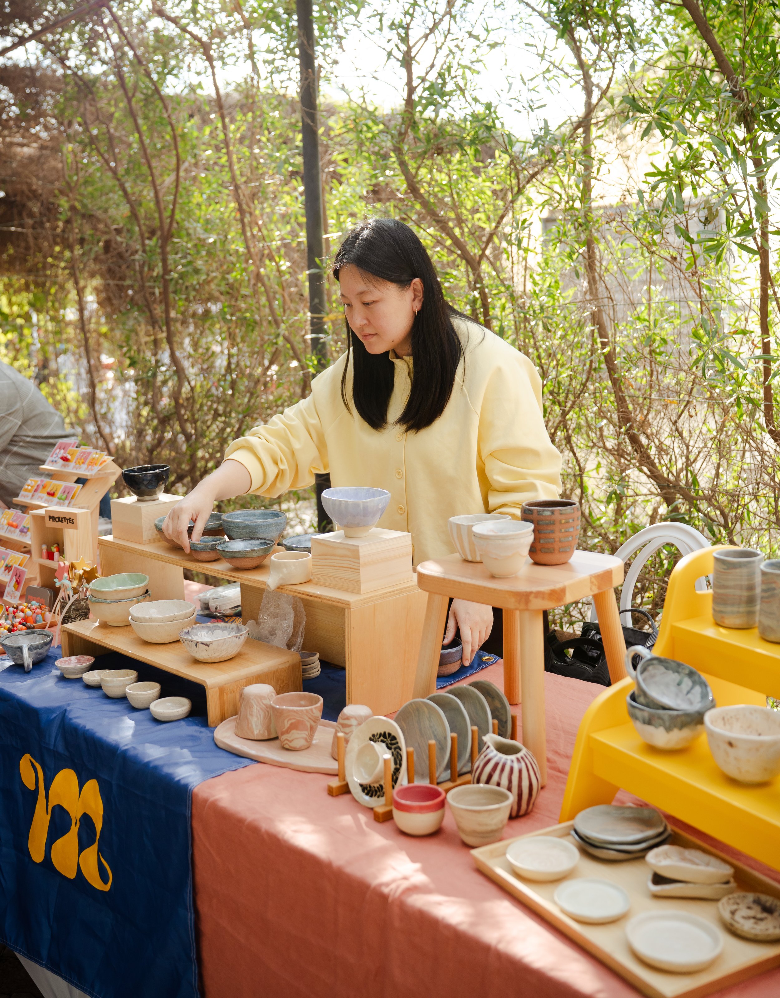 A woman wearing a yellow shirt is selling handmade ceramic pottery at an outdoor craft market stall set up with wooden shelves and tables, surrounded by trees and greenery.