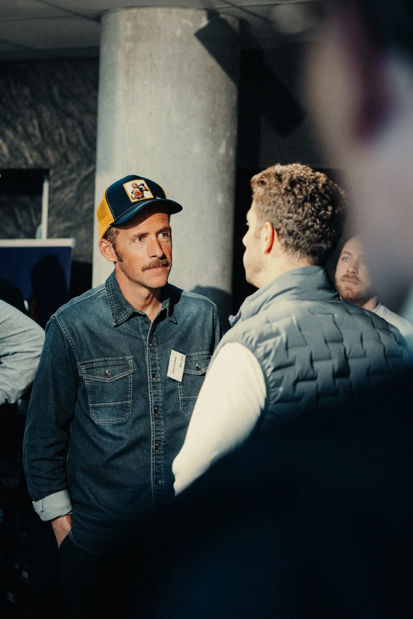 Deux hommes en conversation, l'un porte une casquette colorée et une veste en denim, l'autre porte une veste rembourrée gris foncé. Ils semblent discuter dans un lieu intérieur avec une colonne en béton en arrière-plan.