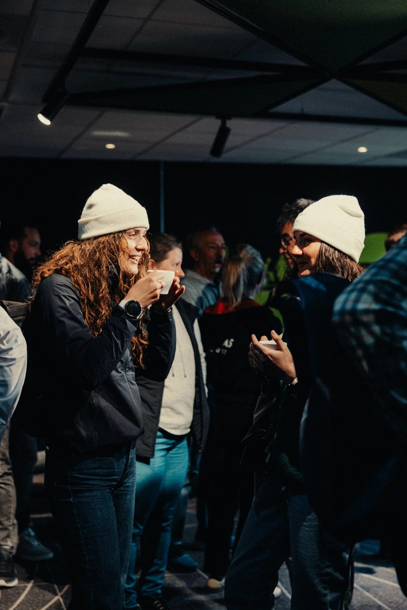 Deux femmes, portant des bonnets blancs, discutent et sourient lors d'un événement en intérieur, entourées d'autres personnes.