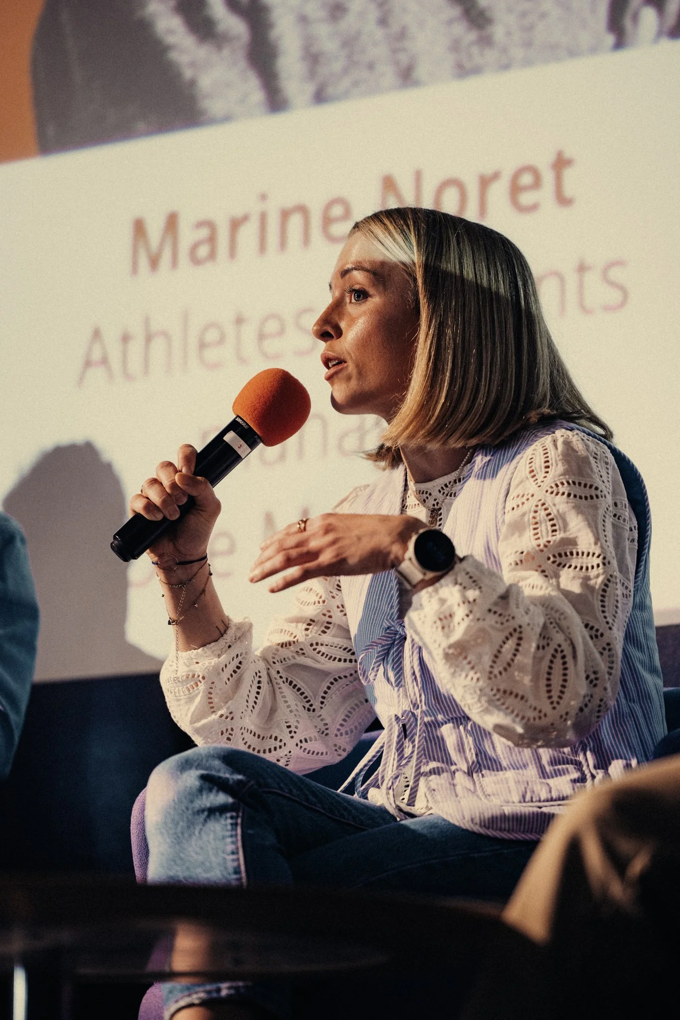 Une femme assise, tenant un microphone orange en train de parler, avec un écran derrière elle affichant du texte en français.