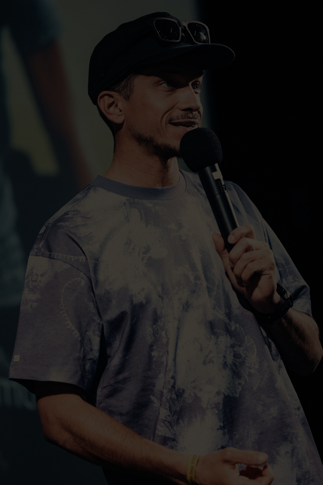 Un homme souriant tient un micro lors d'une conférence ou d'un événement, il porte une casquette noire et un t-shirt à motif