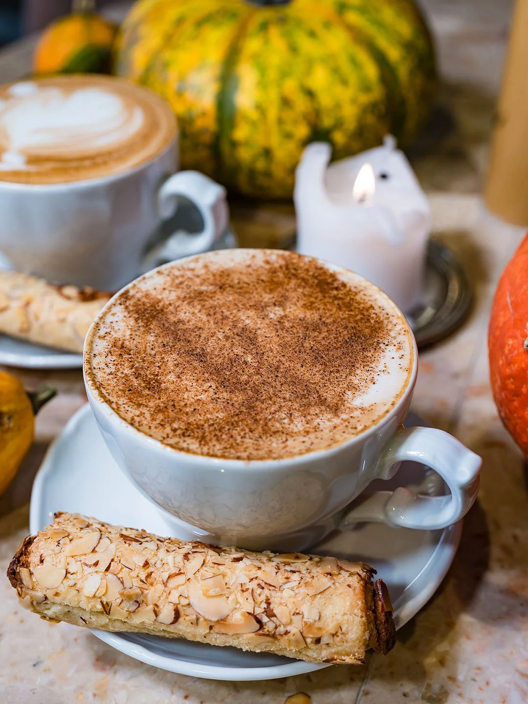 A cup of frothy cappuccino topped with cinnamon, served with an almond biscotti on a saucer, with other fall-themed decorations in the background including pumpkins and a cinnamon-scented candle.