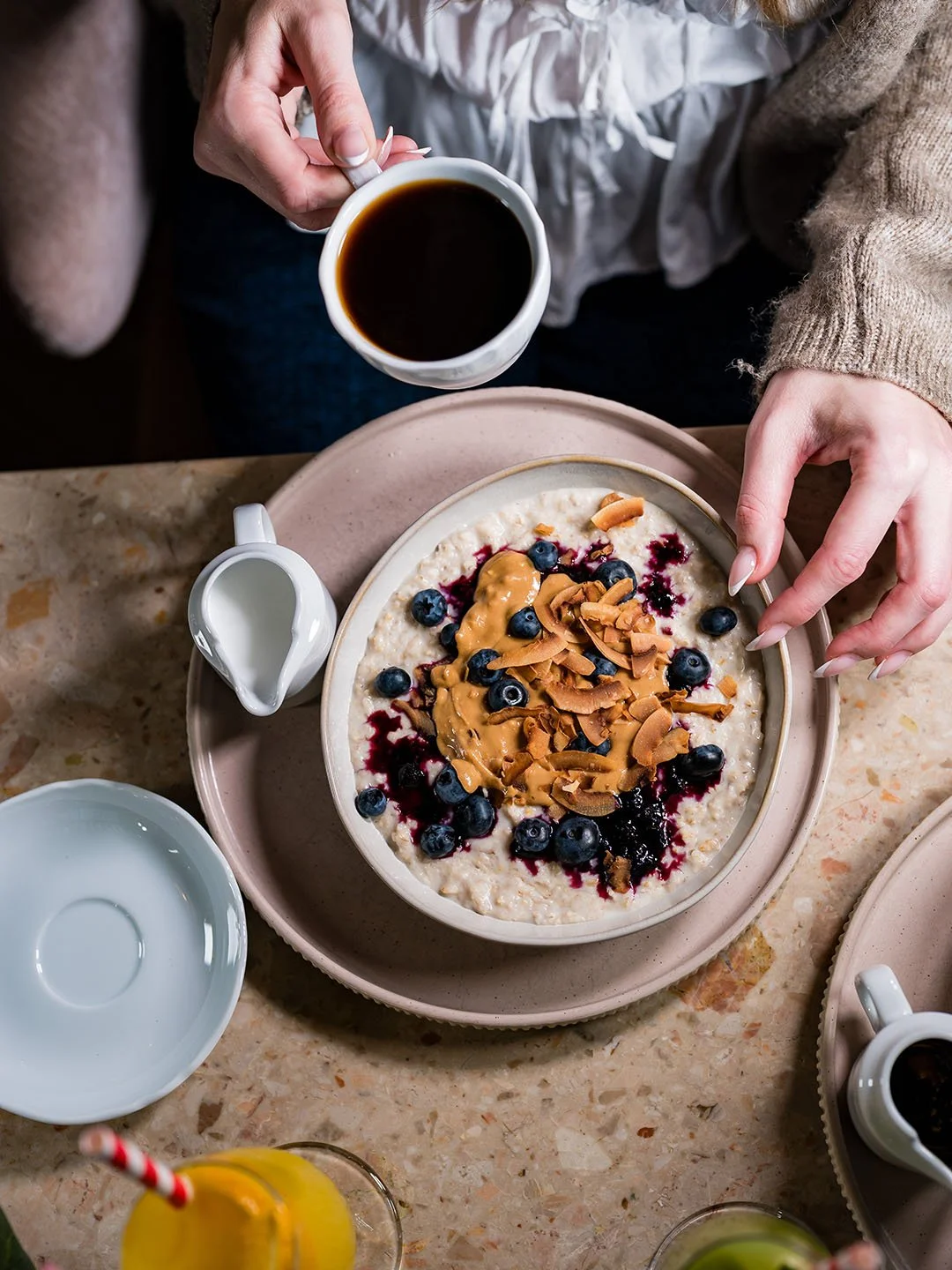 A person holding a cup of coffee with a bowl of oatmeal topped with blueberries, coconut flakes, peanut butter, and berry sauce on a table.