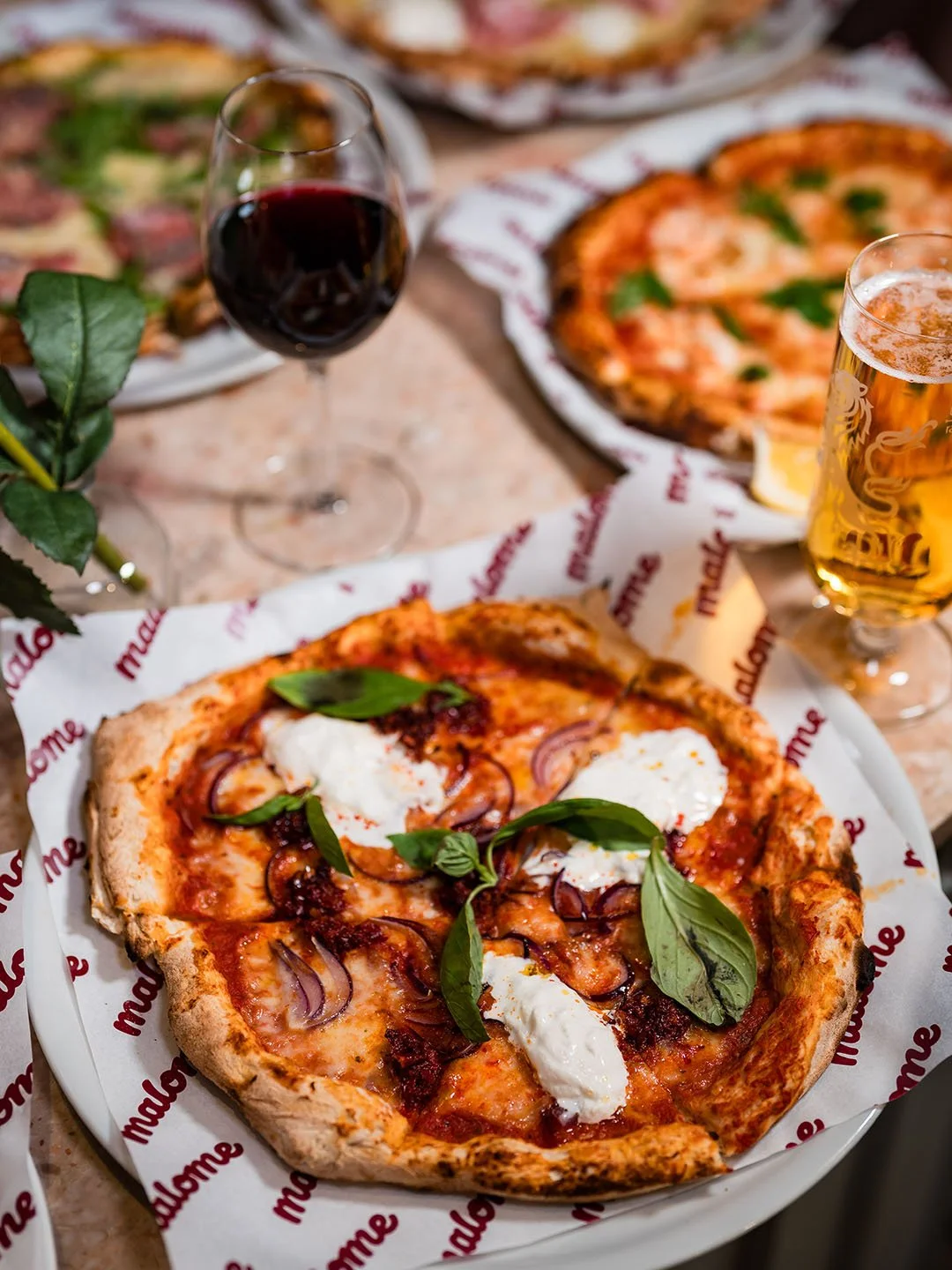 Pizza with mozzarella cheese, basil leaves, and red onions, served on a white plate with Mellow Mushroom branded paper, with glasses of red wine and beer in the background.
