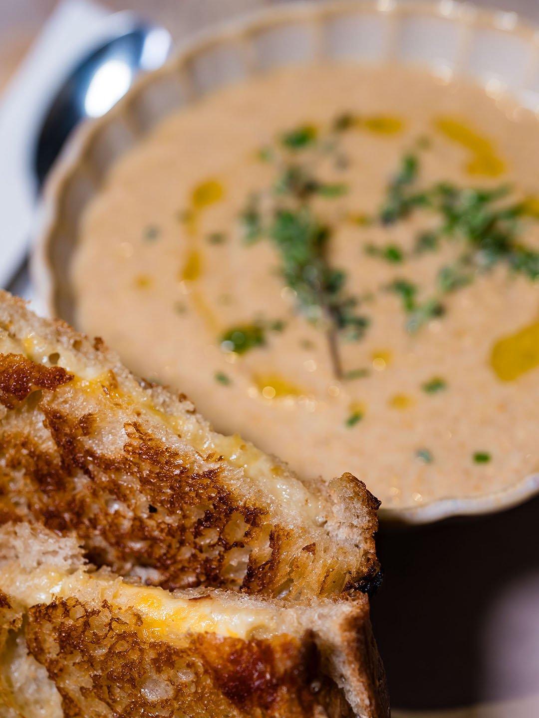 Close-up of a bowl of creamy soup garnished with herbs and drizzled with oil, with a toasted sandwich in the foreground.