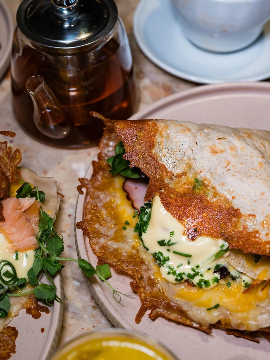 A plate with a breakfast sandwich containing fried egg, ham, cheese, and greens, with a side of another dish topped with smoked salmon and greens, a glass teapot with tea, and cups in the background.
