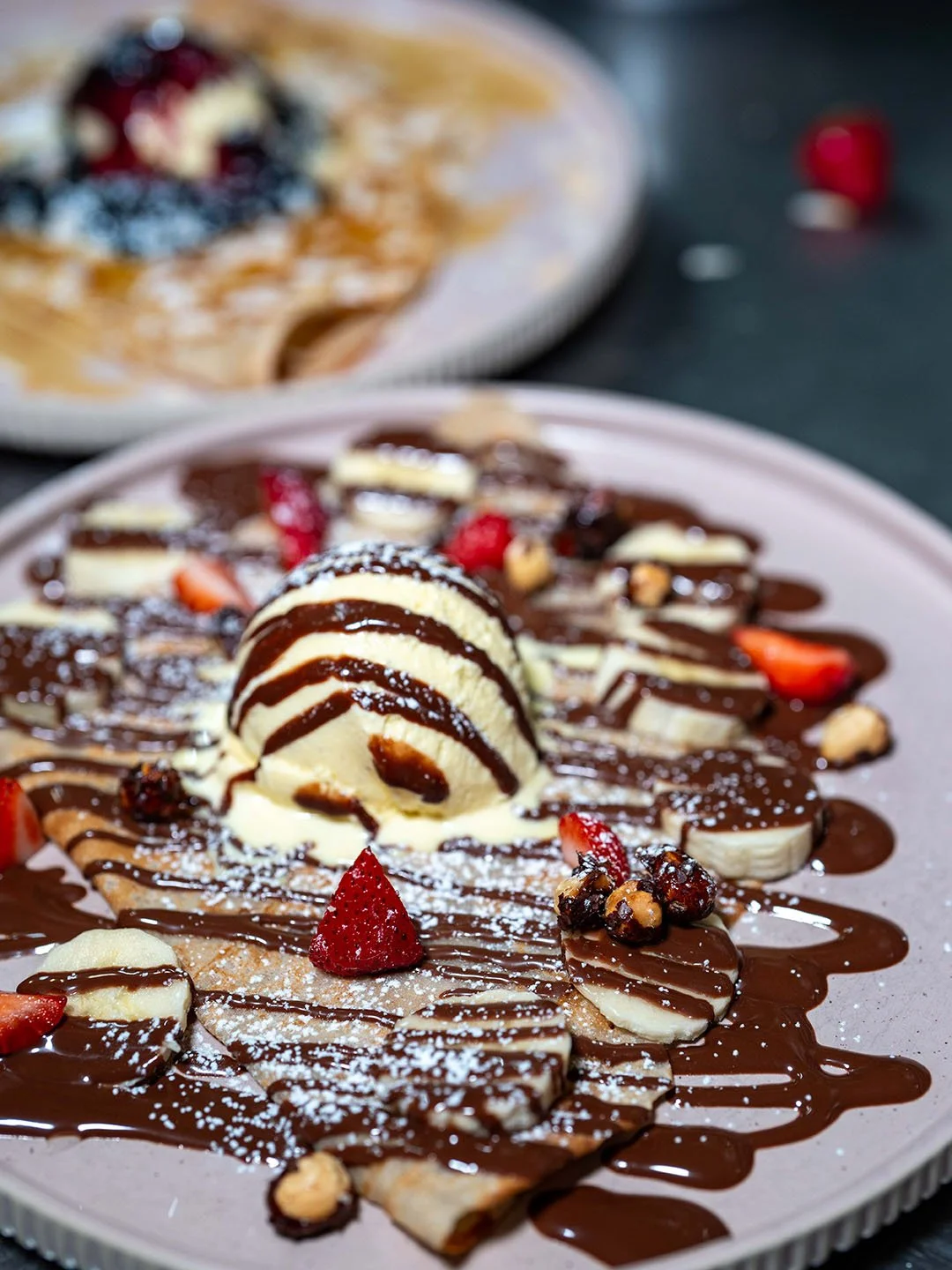 Dessert plate with crepes topped with banana slices, strawberries, mixed berries, chopped nuts, whipped cream, chocolate syrup, and powdered sugar.