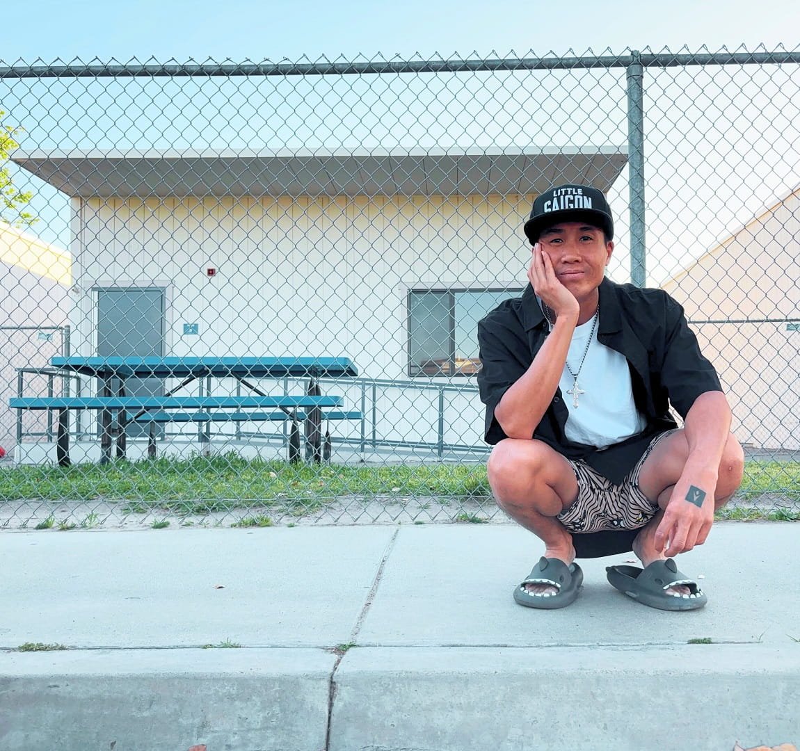 Asian guy named Victor Lam squatting on a sidewalk curb in front of a fence. He is street styled wearing a black cap with the words 'Little Saigon', a black open shirt, white t-shirt, striped Badtz Maru shorts, shark slides, and a cross necklace.