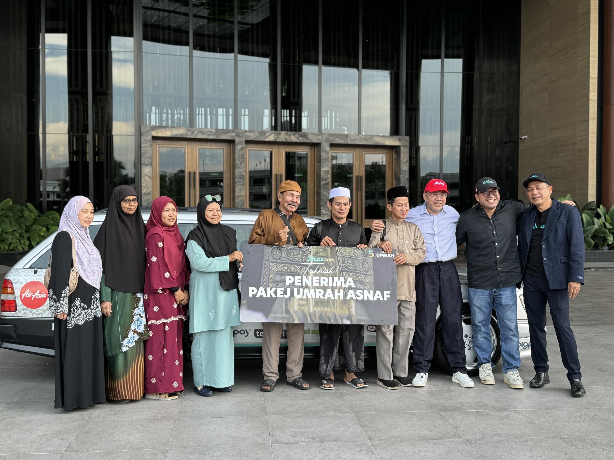 A group of diverse people standing in front of a building, holding a large sign that reads "Penerima Pakej Umrah Asnaf". There is a car behind them and the building has large glass windows.