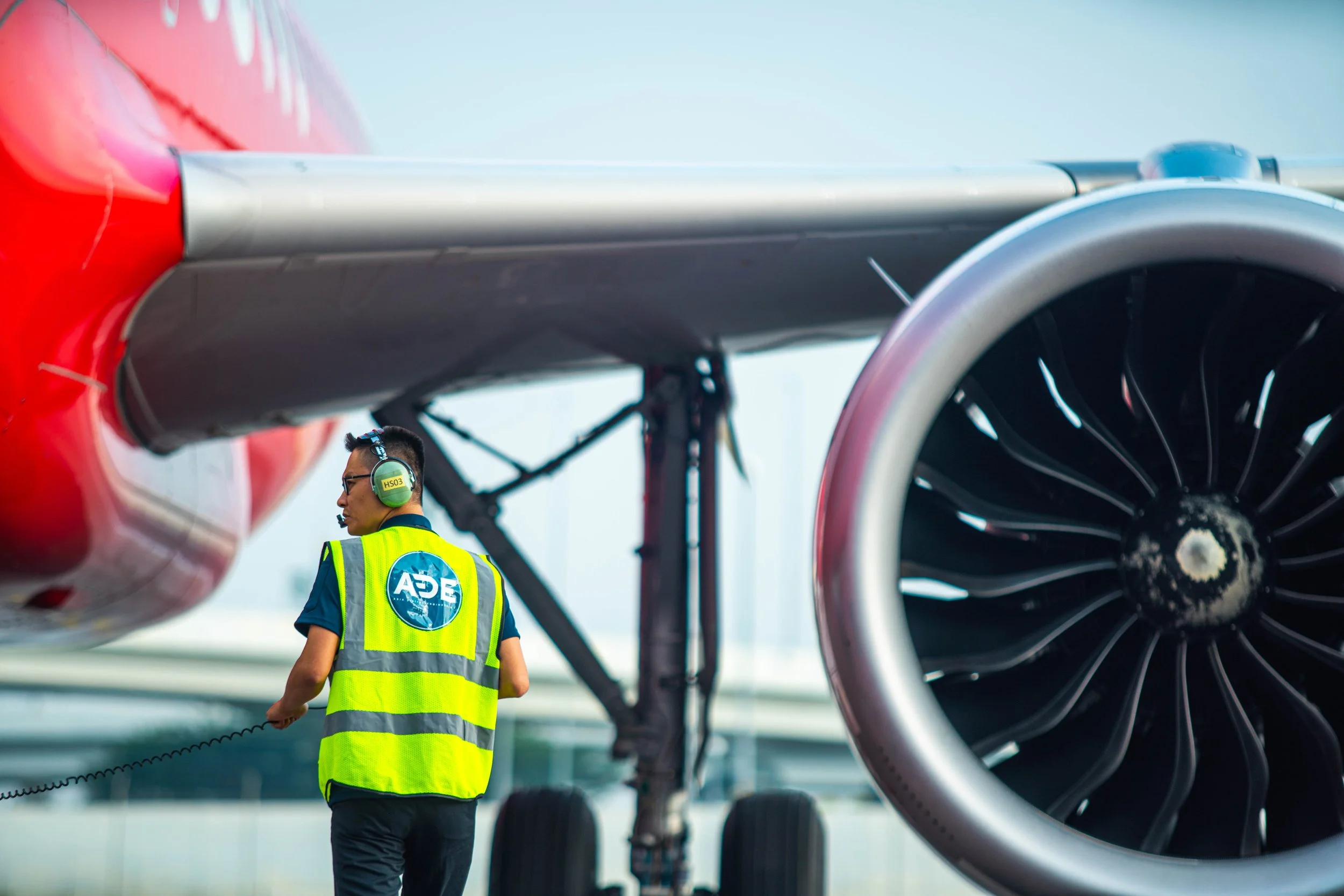 An aircraft ground crew member wearing a high-visibility vest with the ADE logo and a headset, walking near a parked jet engine on an airfield.