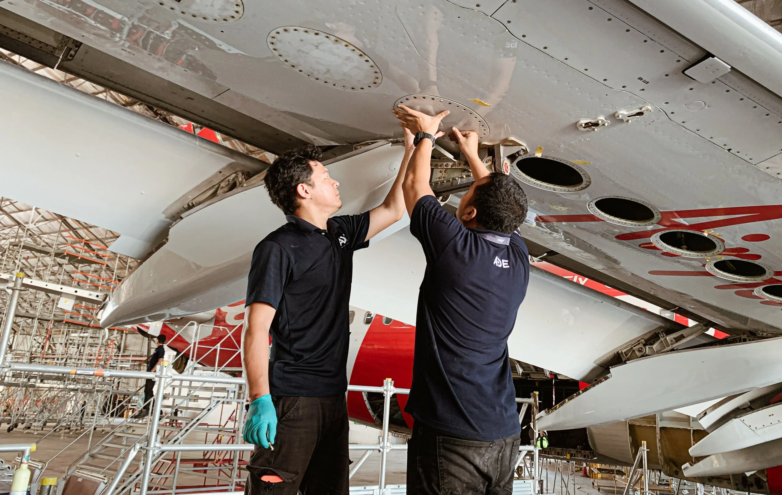 Two men inspect and work on the underside of an aircraft in a large aircraft maintenance hangar. They're using tools and wearing uniforms.