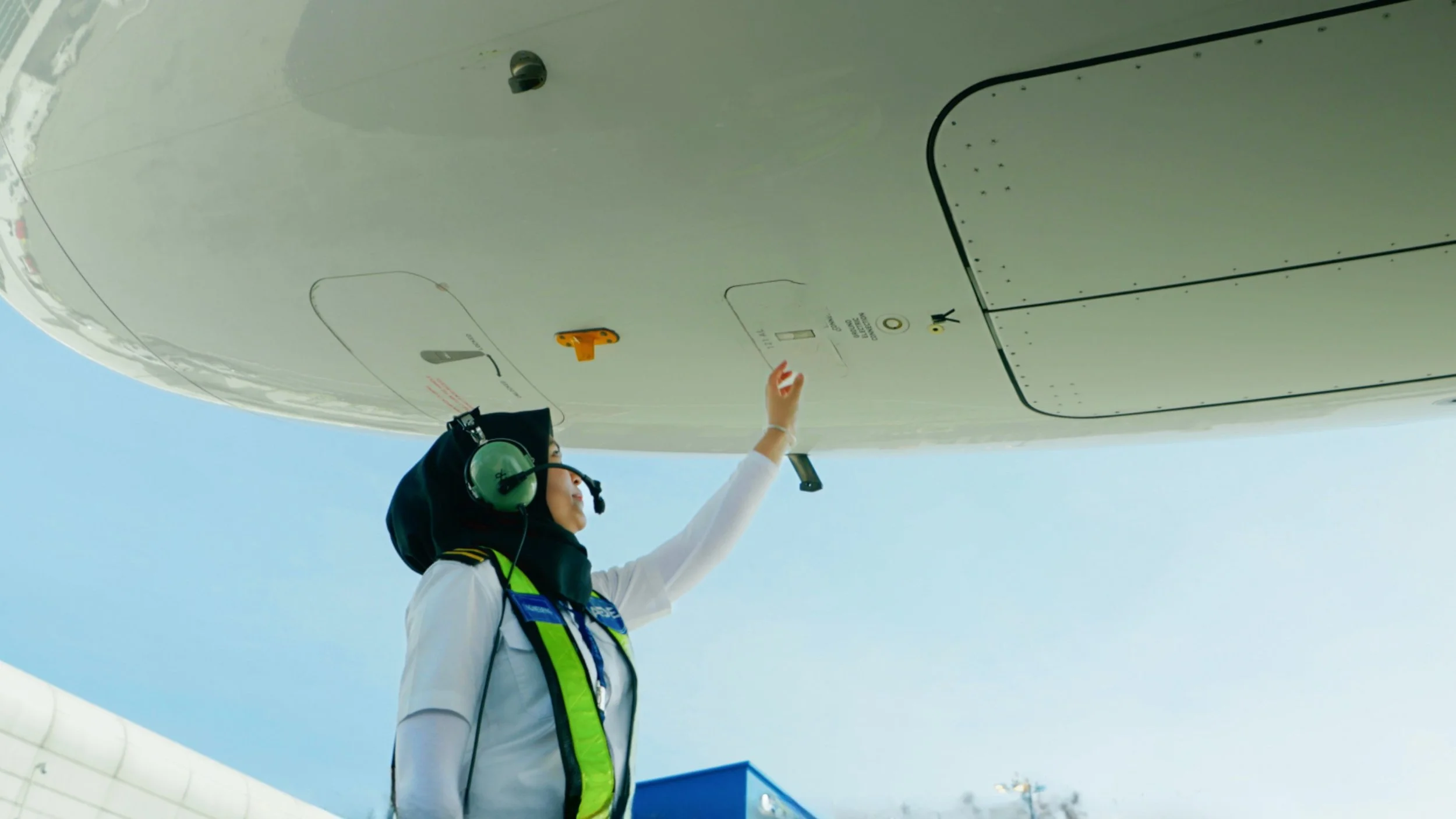 A woman in aviation headset and safety vest reaching up to touch a part of an aircraft's undercarriage.