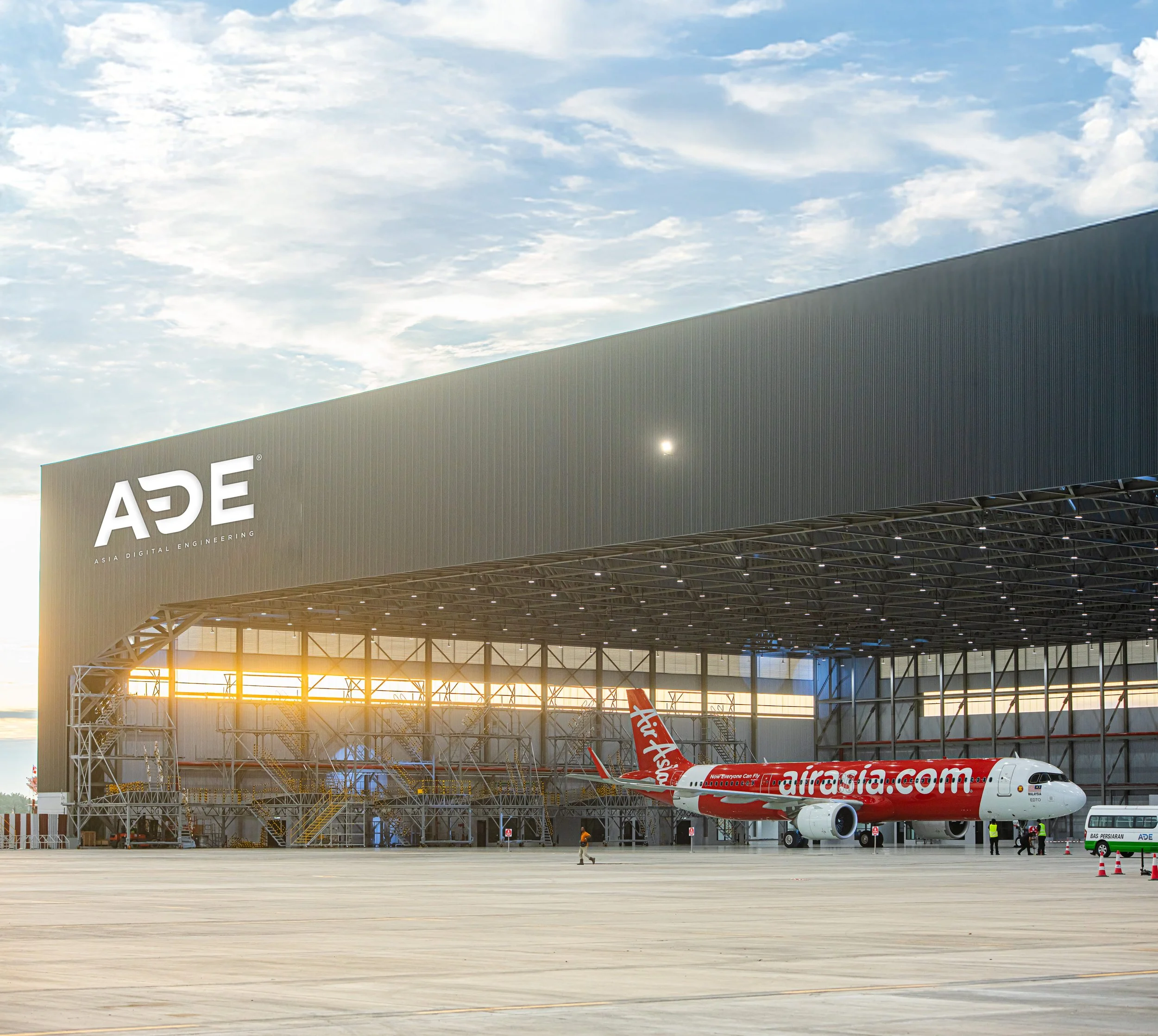 An airplane on the tarmac of an airport with a large building in the background. The airplane is from airasia.com and is parked near a jet bridge. The sky is partly cloudy and the scene is illuminated by sunlight.