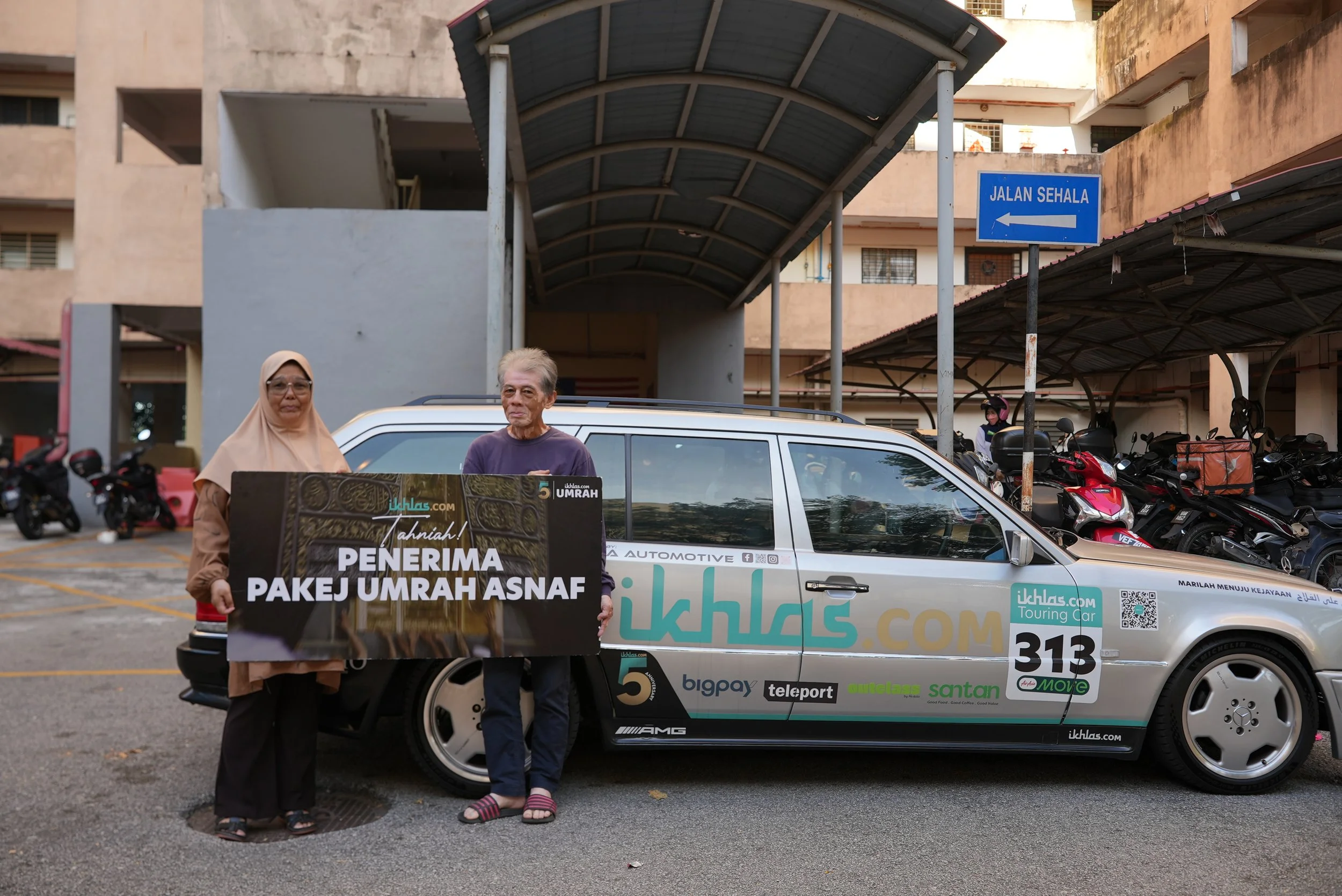 An older woman and a man standing in front of a silver car with Uber and ikhlas.com branding, holding a banner that reads "Tahniah! Penerima Pakej Umrah Asnaf" in a parking lot with motorcycles and apartment buildings in the background, under a covered walkway and a blue street sign indicating "Jalan Sehala" to the left.