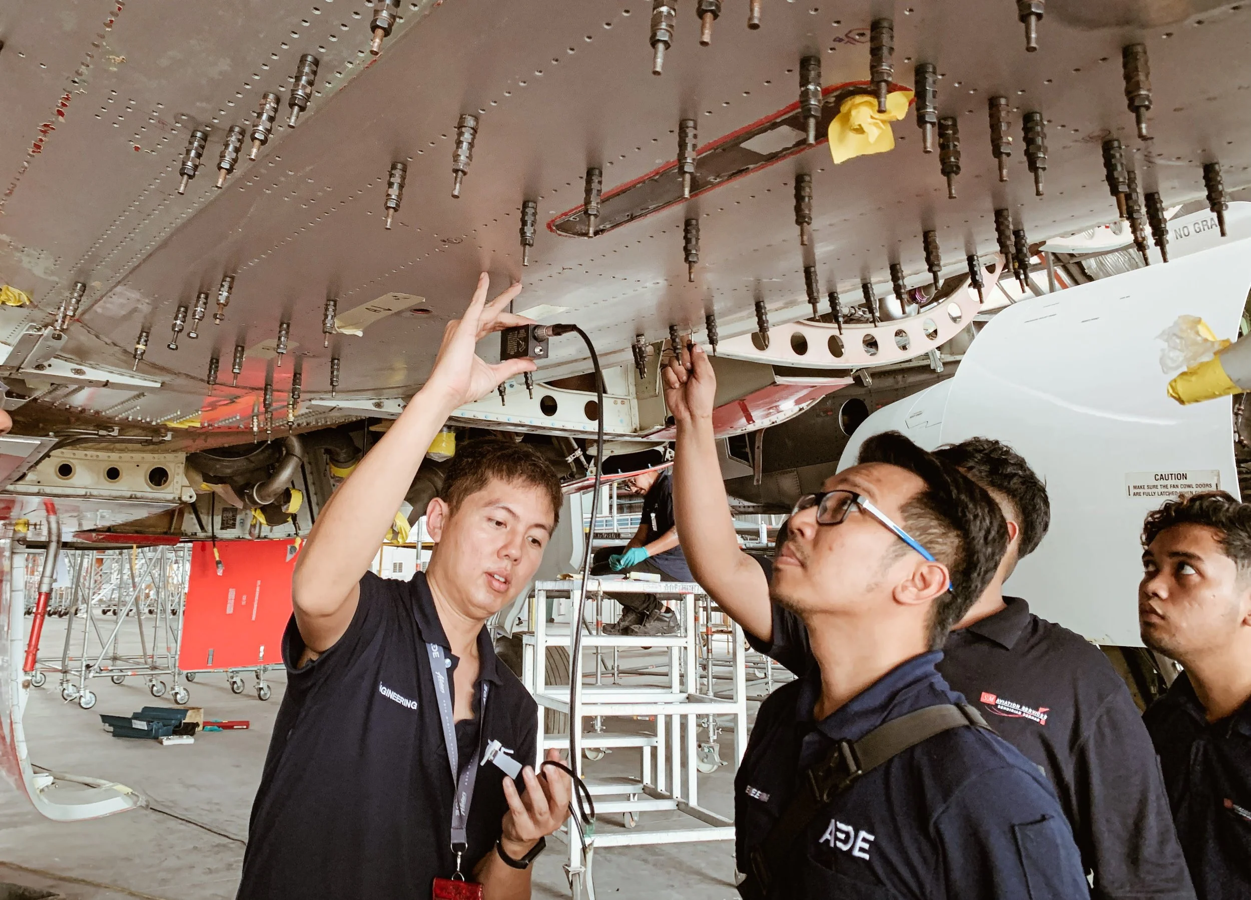 Group of people working on aircraft maintenance, inspecting and handling tools underneath a large aircraft fuselage with various panels and fasteners visible.