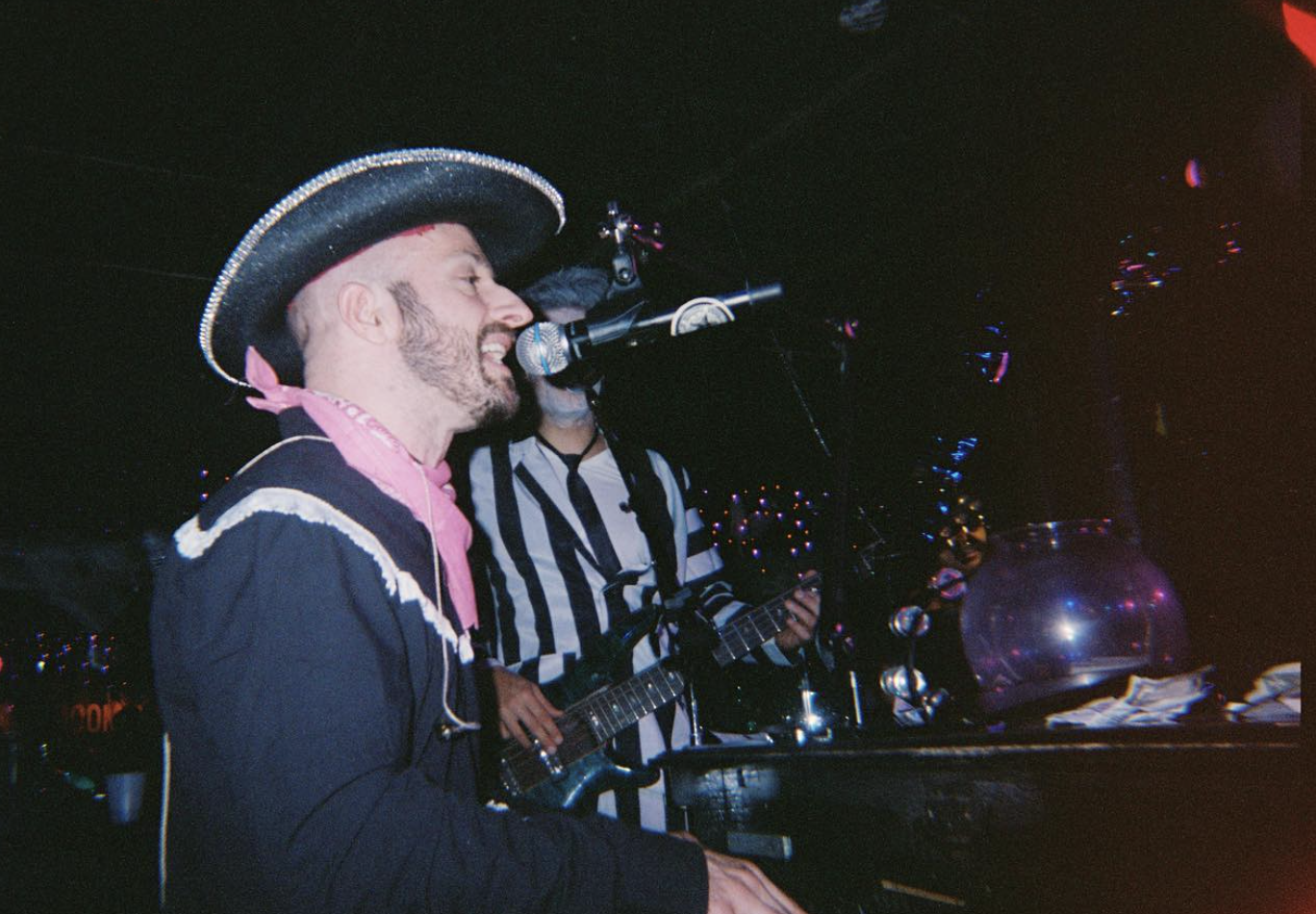 A man wearing a traditional Mexican sombrero and pink bandana singing into a microphone, with another person playing guitar in striped outfit in the background, performing on stage at a lively event.