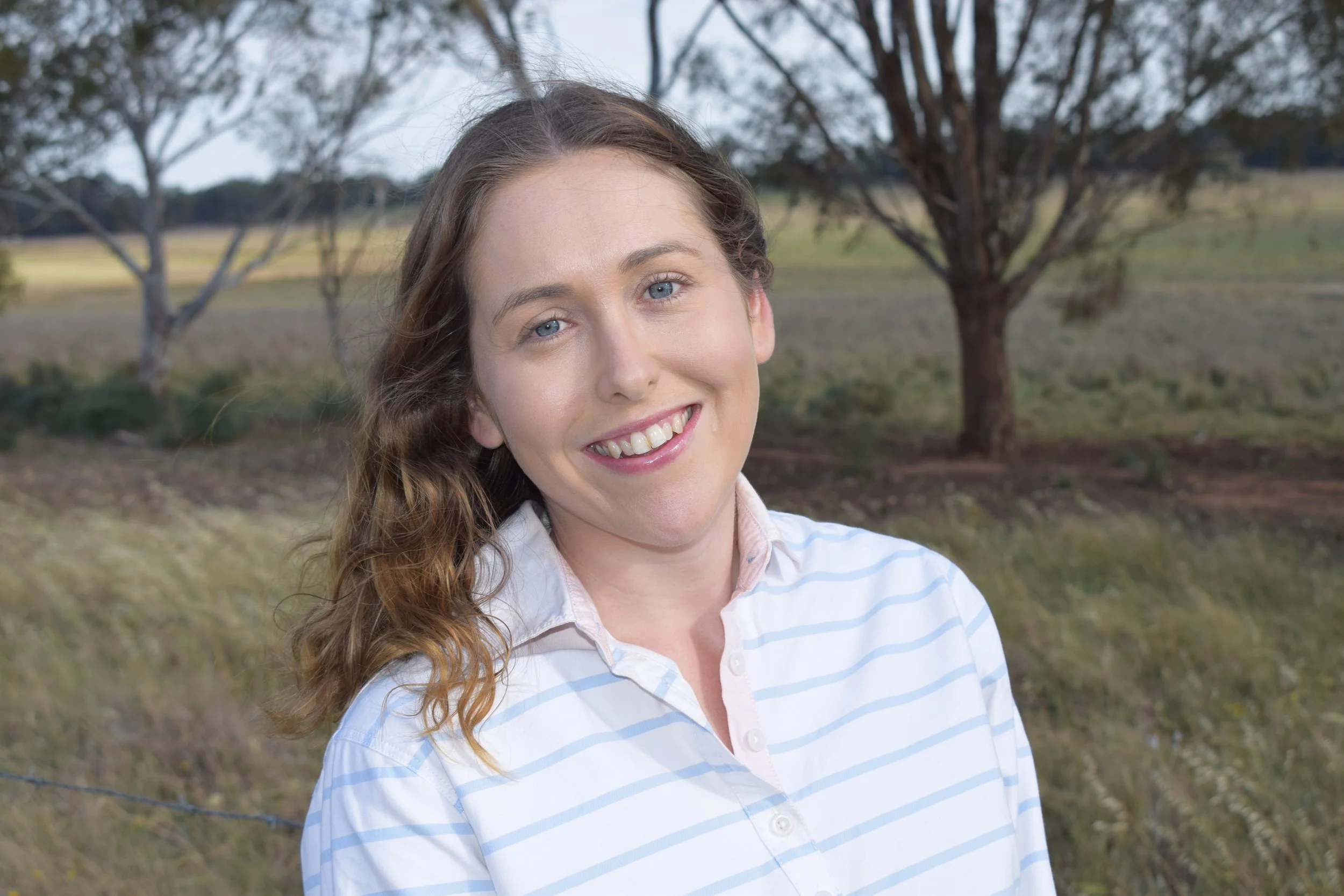 A smiling woman with light skin, wavy brown hair, and blue eyes, wearing a white and light blue striped shirt, outdoors with trees and grassy paddock in the background.