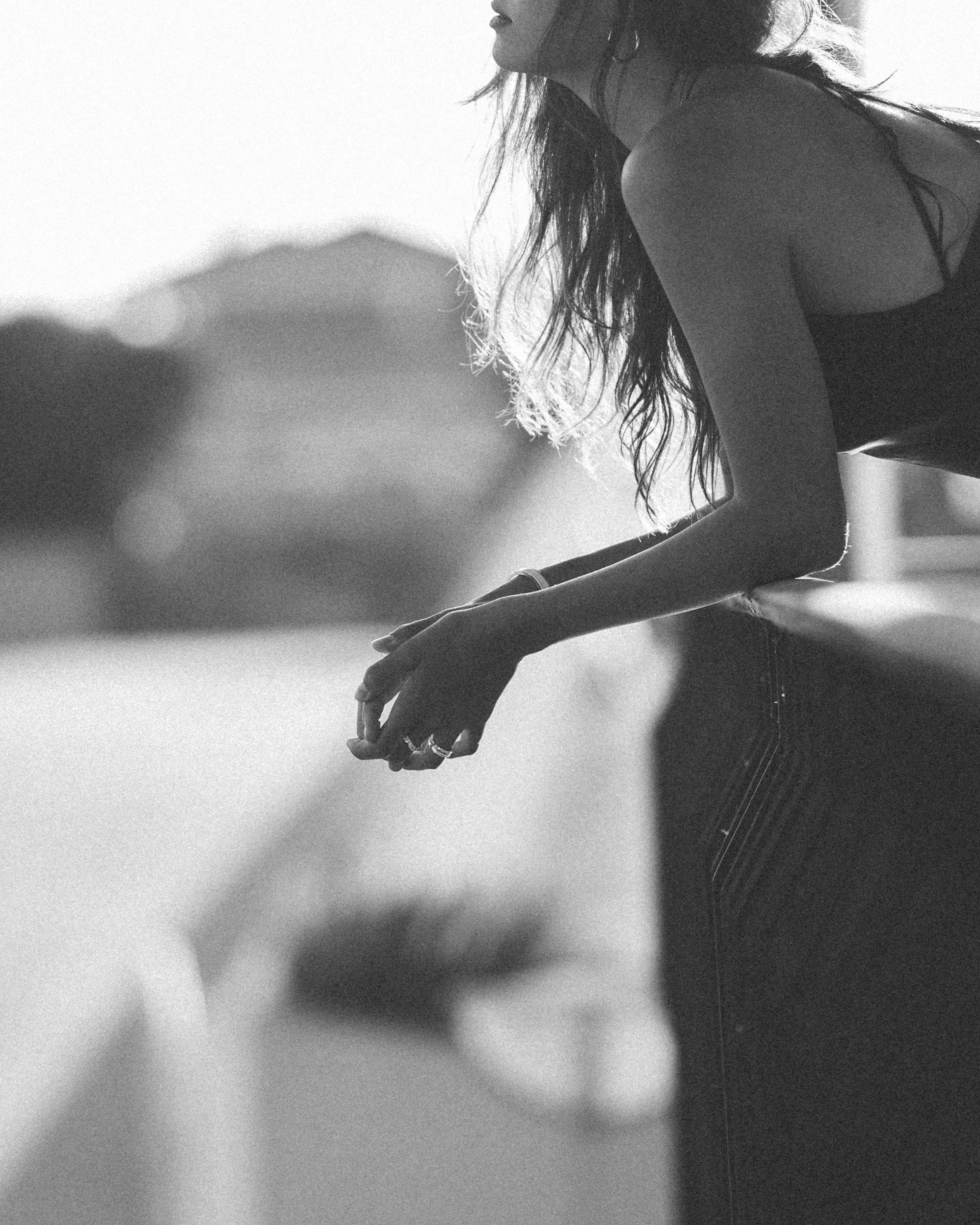 A black-and-white photo of a woman leaning on a railing, with her face turned to the side and her hair hanging over her shoulder, outdoors during daylight.