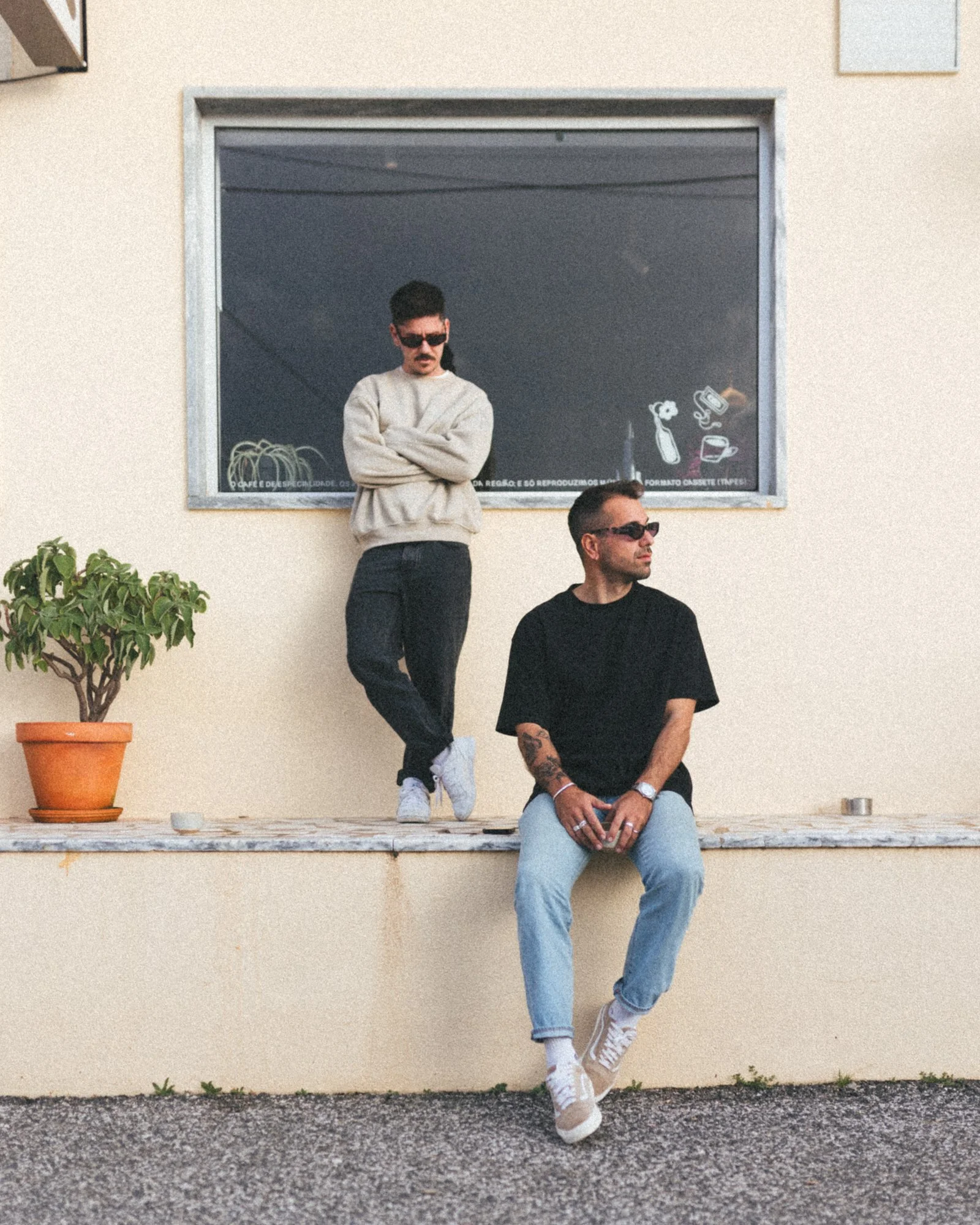 Two young men wearing sunglasses pose outside a building; one sits on a ledge with crossed legs, wearing a black t-shirt and light jeans, while the other stands with arms crossed, wearing a beige hoodie and dark jeans.