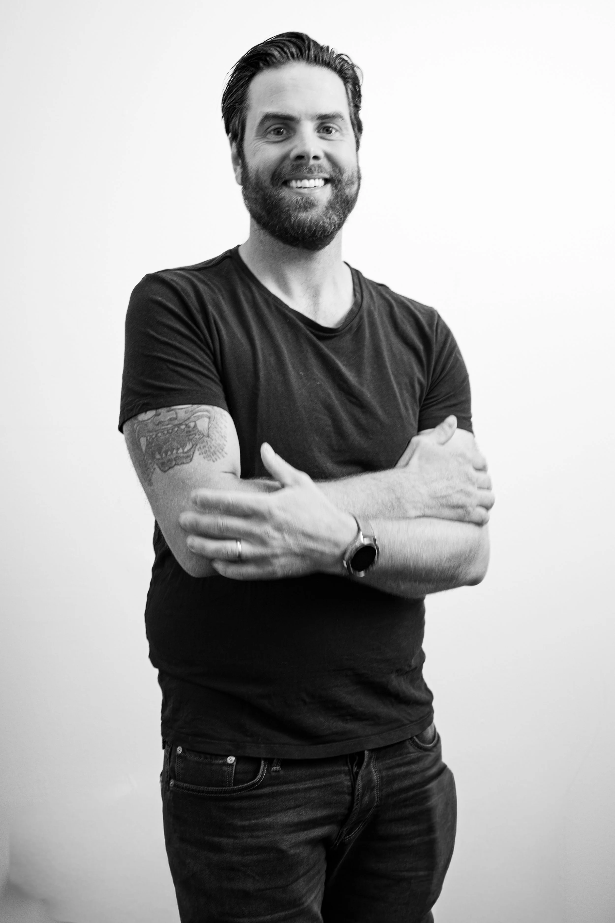 En man med mörkt hår, skägg och tatuering på armen, står mot en vit bakgrund, bär svart T-shirt och jeans, med armar korsade och ler.