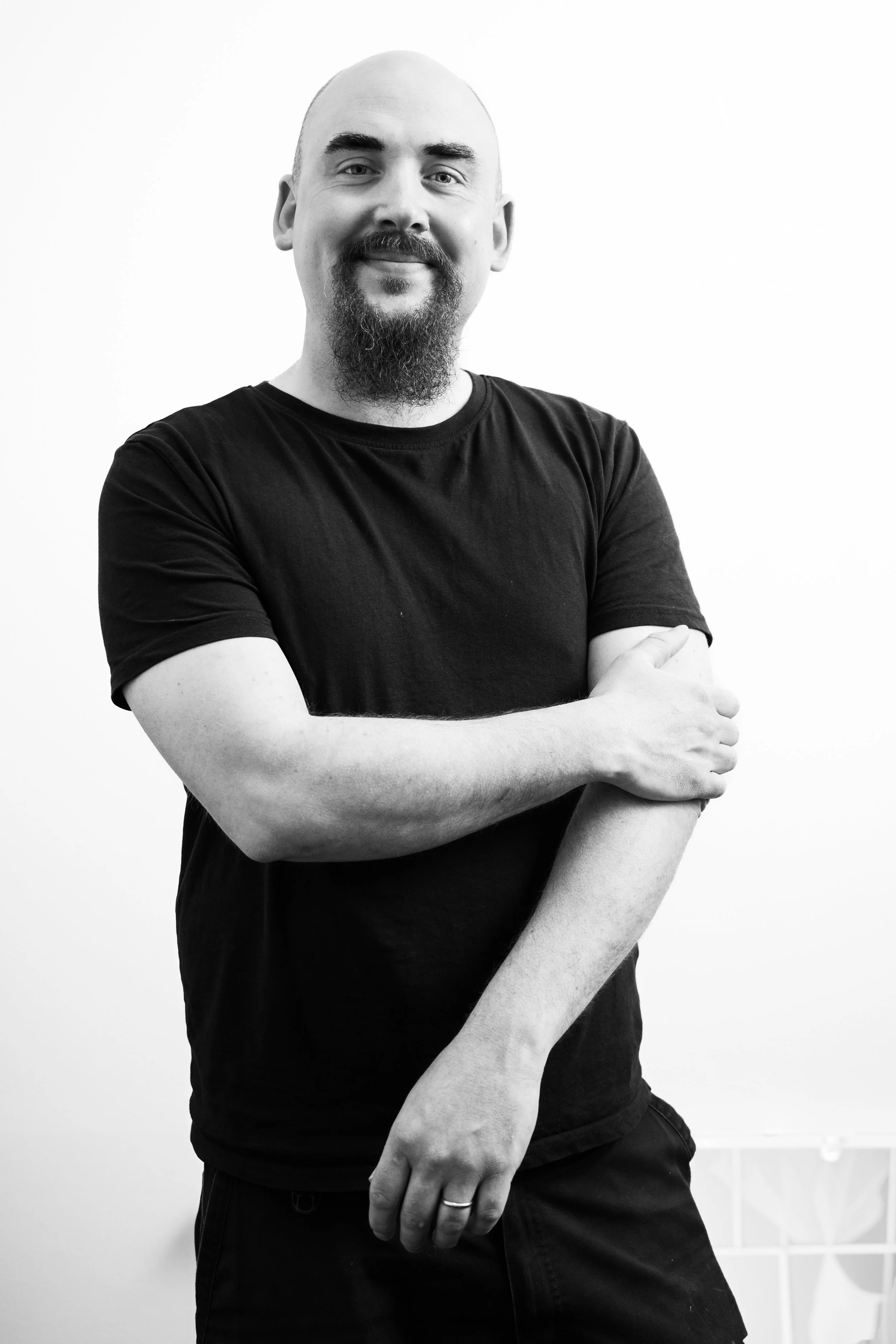 Svartvit bild av en man med skägg och mustasch, bär svart T-shirt, korsar armarna framför en vit vägg.