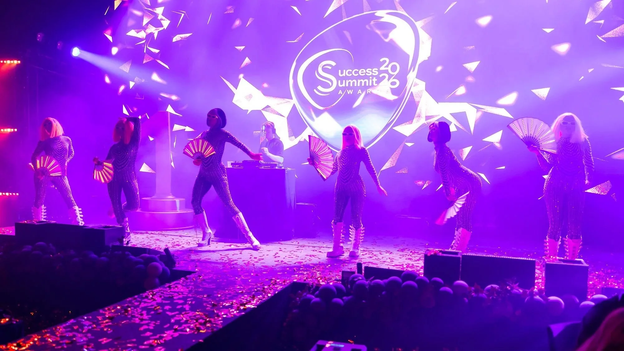 På scenen är en grupp damer i skinnende kläder som dansar med fans i händerna. Bakom dem projiceras en ljusbild för Success Summit 2026. Scenbelysningen är lila och konfetti ligger på golvet.