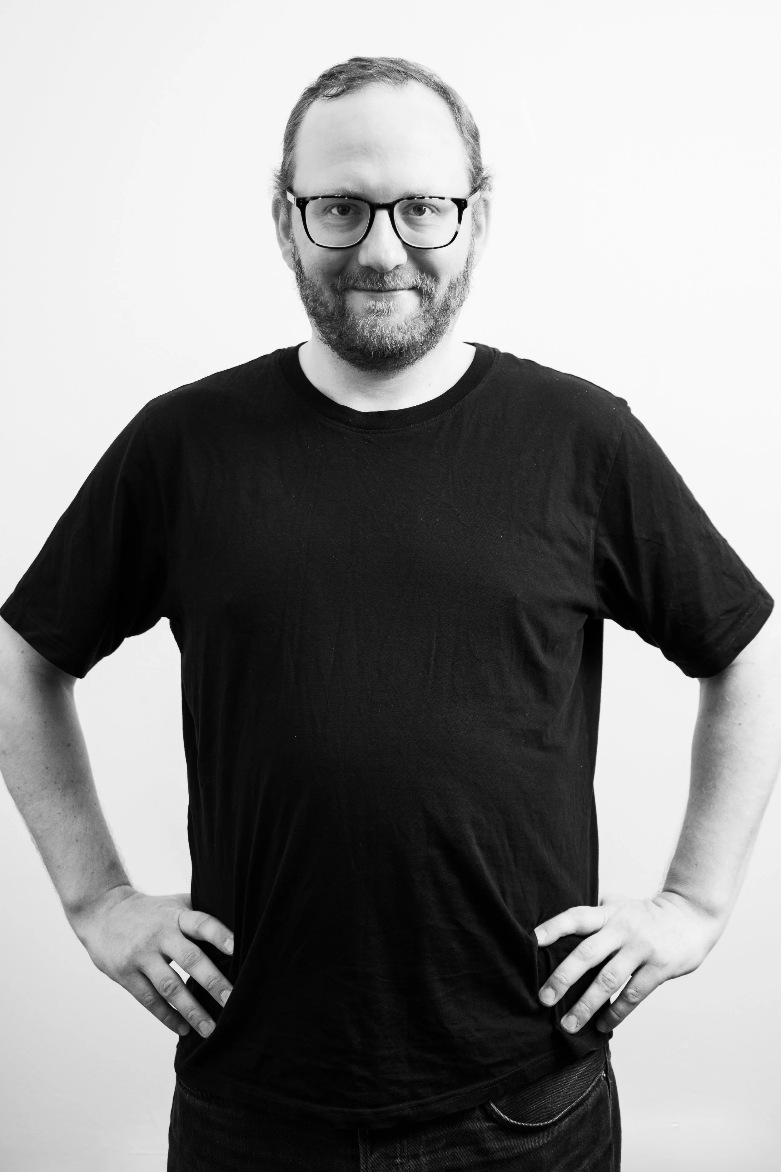 Svartvitt foto av en man med glasögon, skägg och kort hår, som står med händerna på höfterna mot en vit bakgrund.