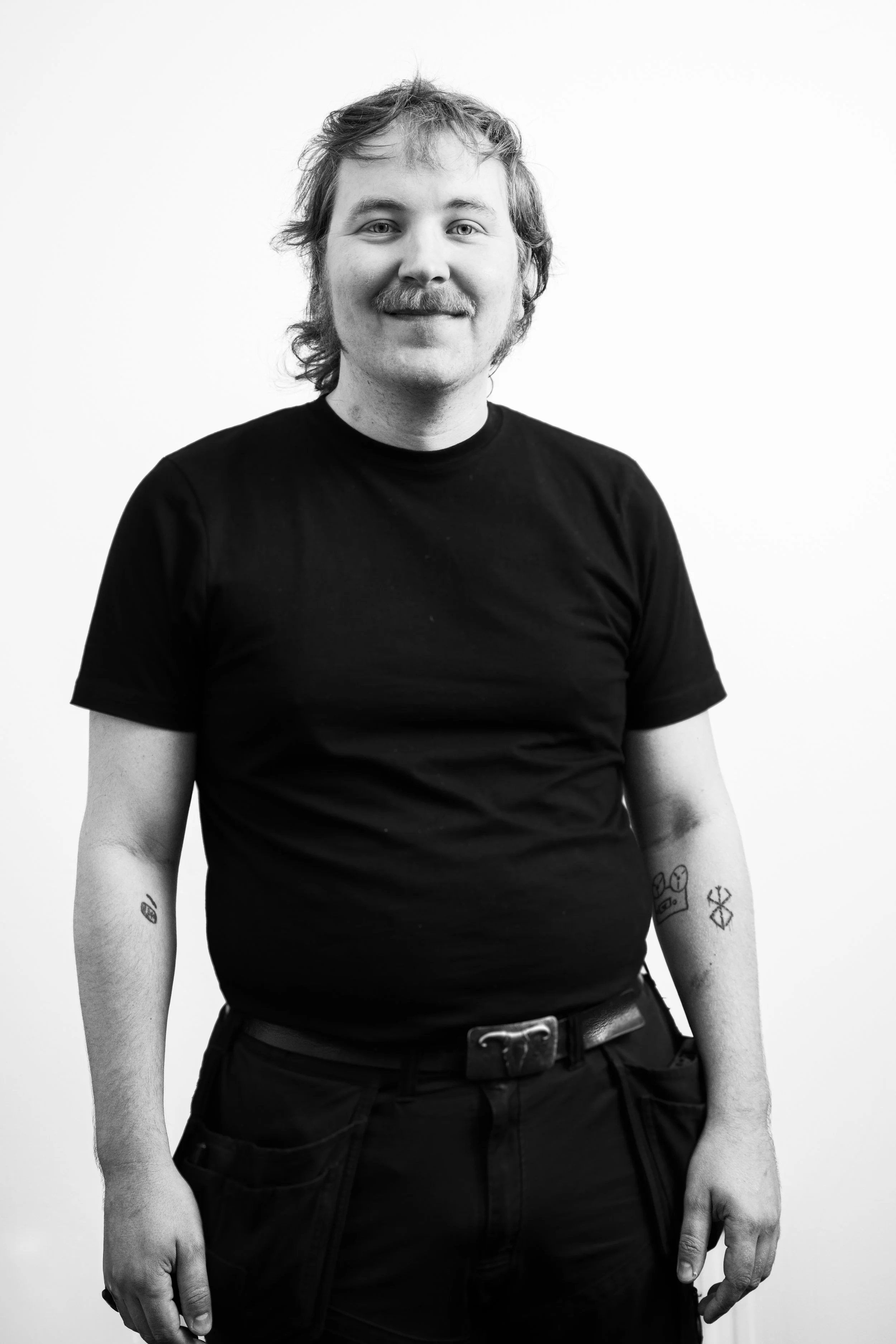 Svartvit bild av en man som står mot en vit vägg. Han bär en svart T-shirt och har tatueringar på båda underarmarna. Han har mellanlångt, lockigt hår och skägg. Han ler med stängd mun och tittar mot kameran.