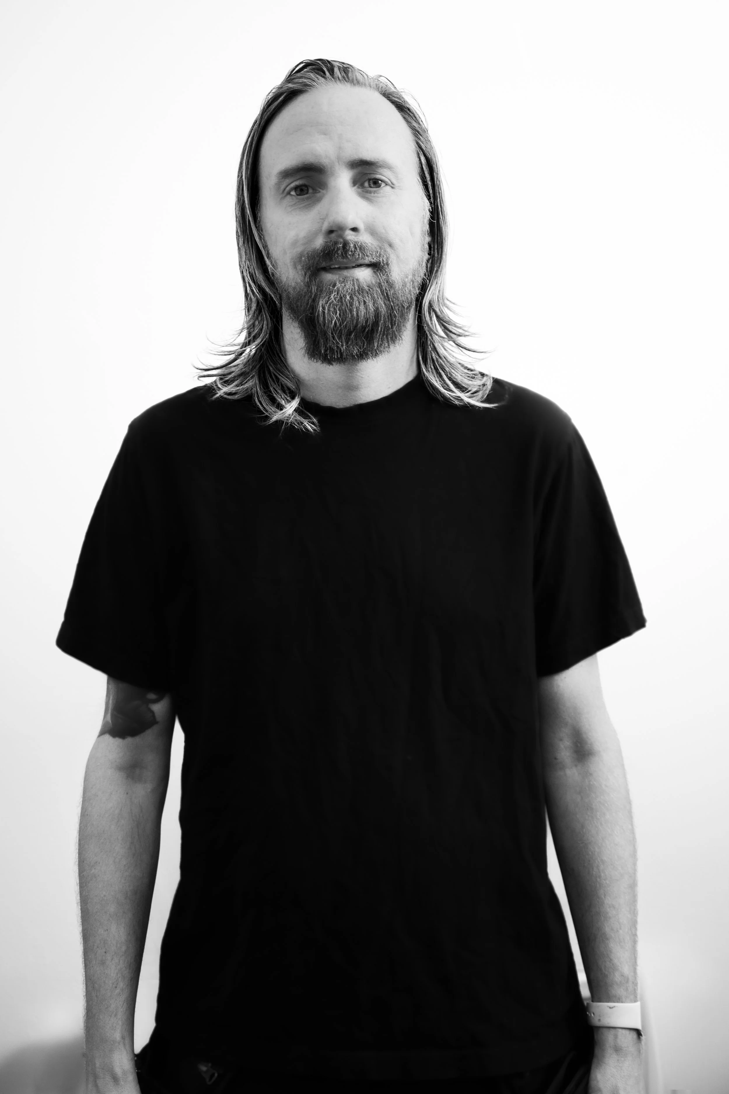 Svartvit bild av en man med långt hår och skägg, bär en svart T-shirt, står framför en vit vägg.