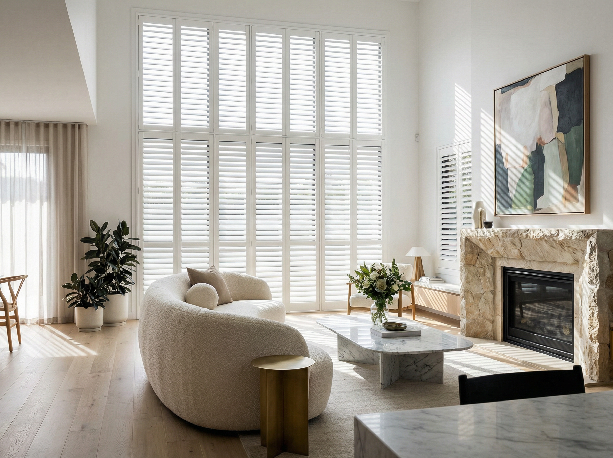 Bright living room with large window covered in white shutters, a stone fireplace, white curved sofa, marble coffee table, potted plant, and colorful abstract artwork.