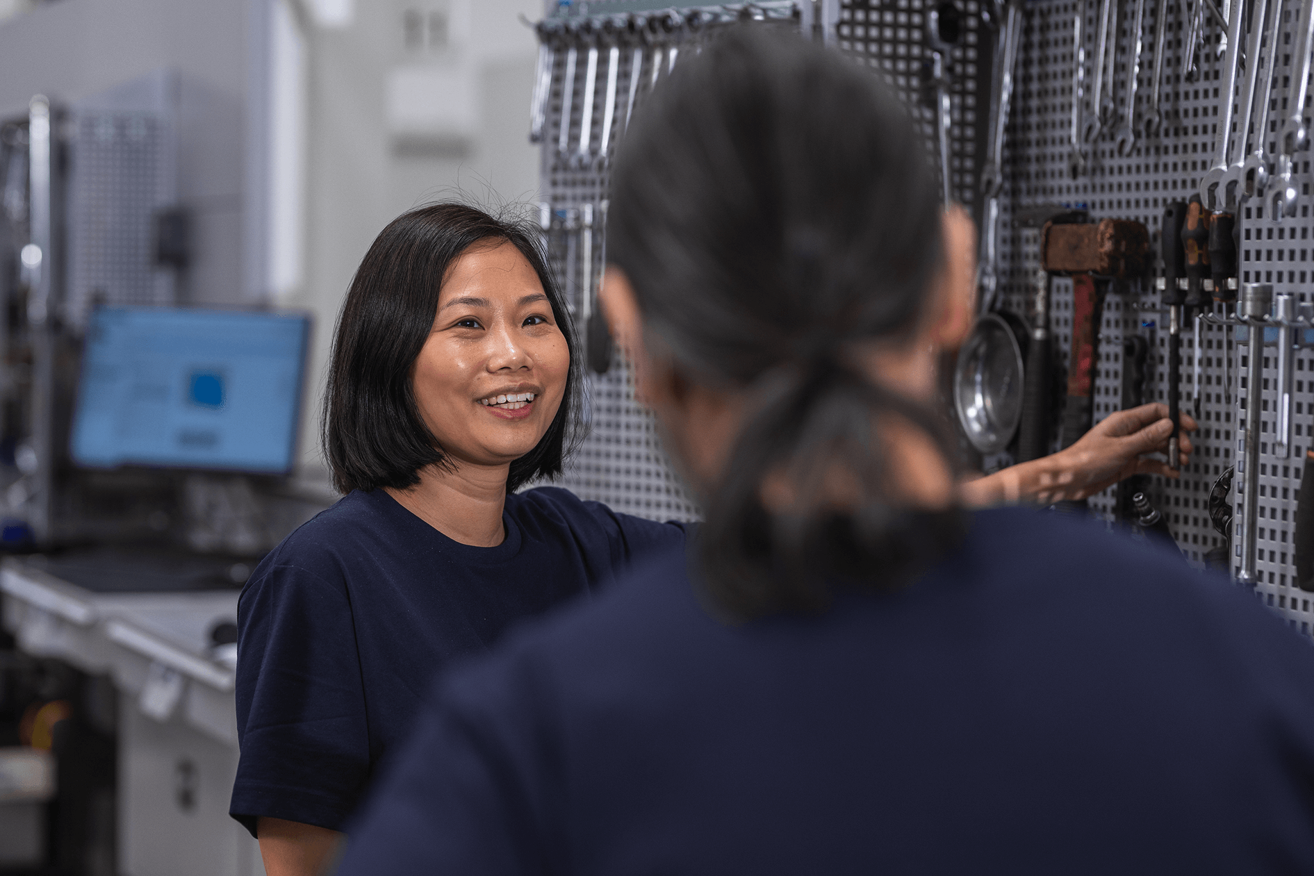 Two Volvo technicians in a workshop or manufacturing setting, one smiling and looking at the other, with tools hanging on a pegboard in the background.