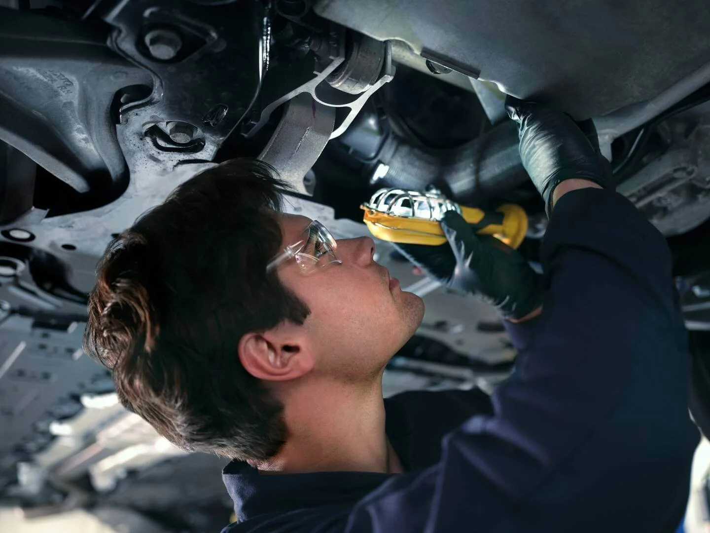 A graduate of the Volvo Apprentice Technician Program wearing safety goggles and gloves is repairing a car underneath the vehicle, using a screwdriver.