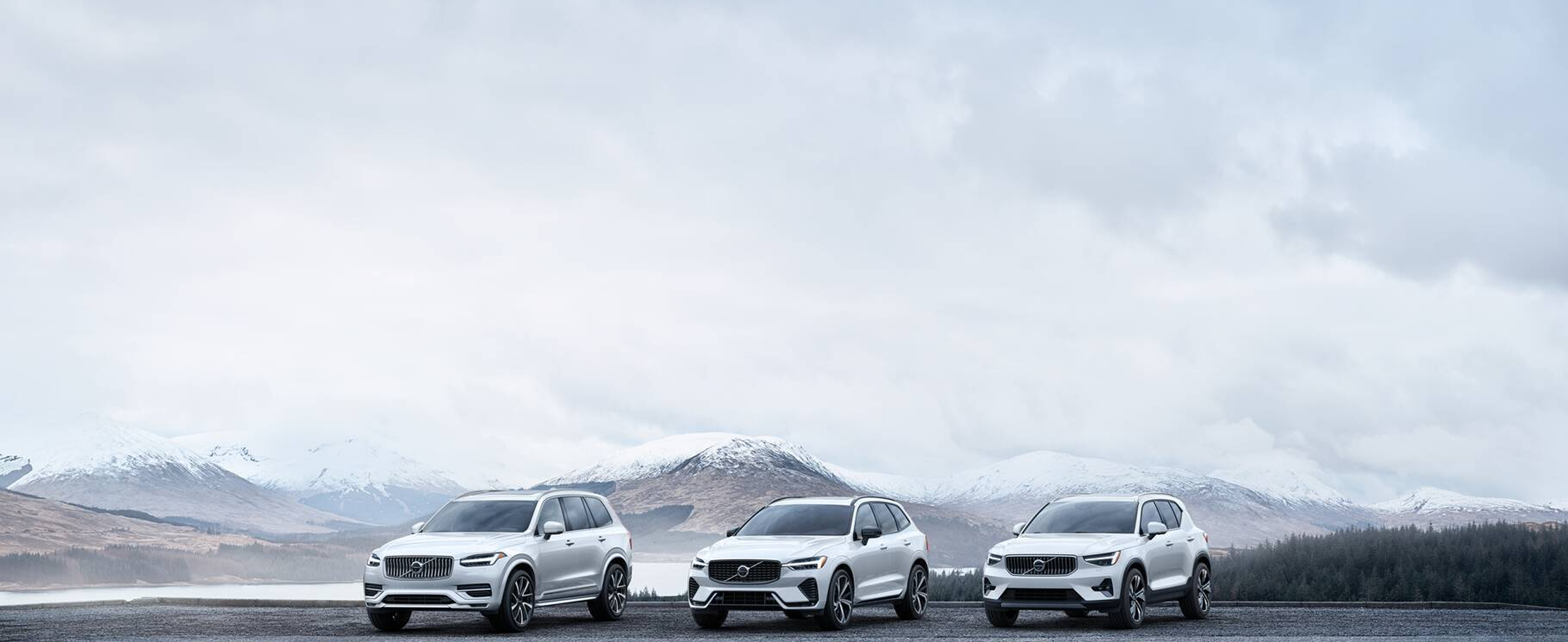 Three white Volvo SUVs parked on a wet road with snow-capped mountains and cloudy sky in the background.