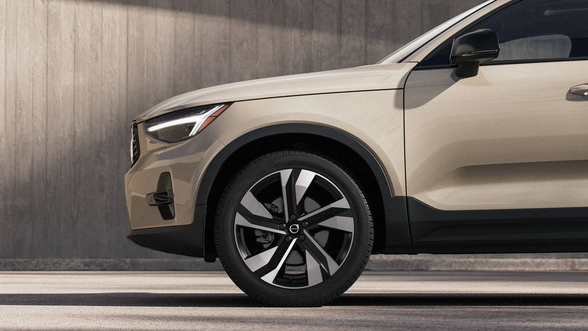 Close-up of the front part of a beige electric Volvo car, focusing on the front wheel and headlight, with a plain wall in the background.