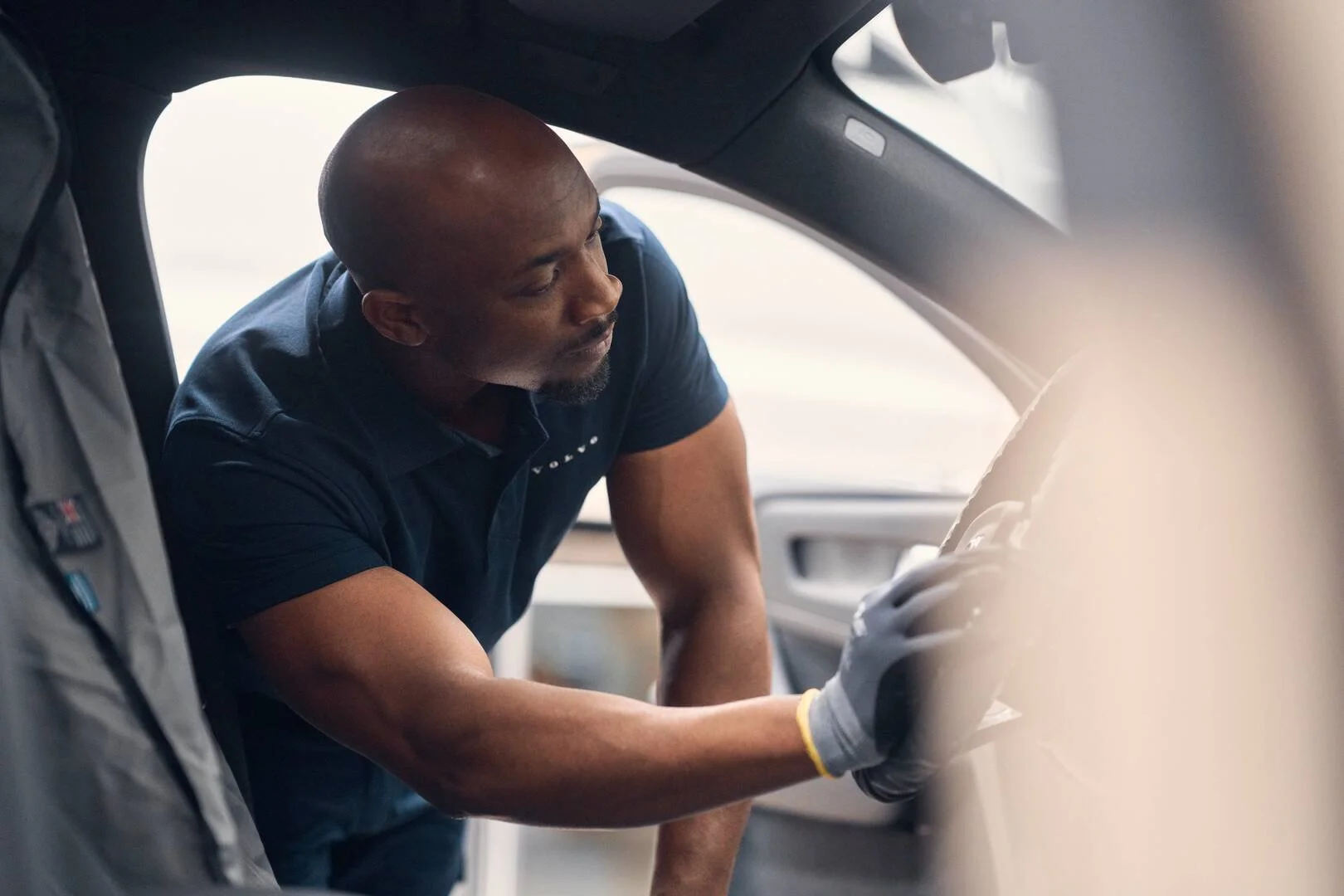 A graduate of the Volvo Apprentice Technician Program wearing a blue uniform and gray gloves is leaning into a vehicle's interior, possibly working on or inspecting the vehicle.