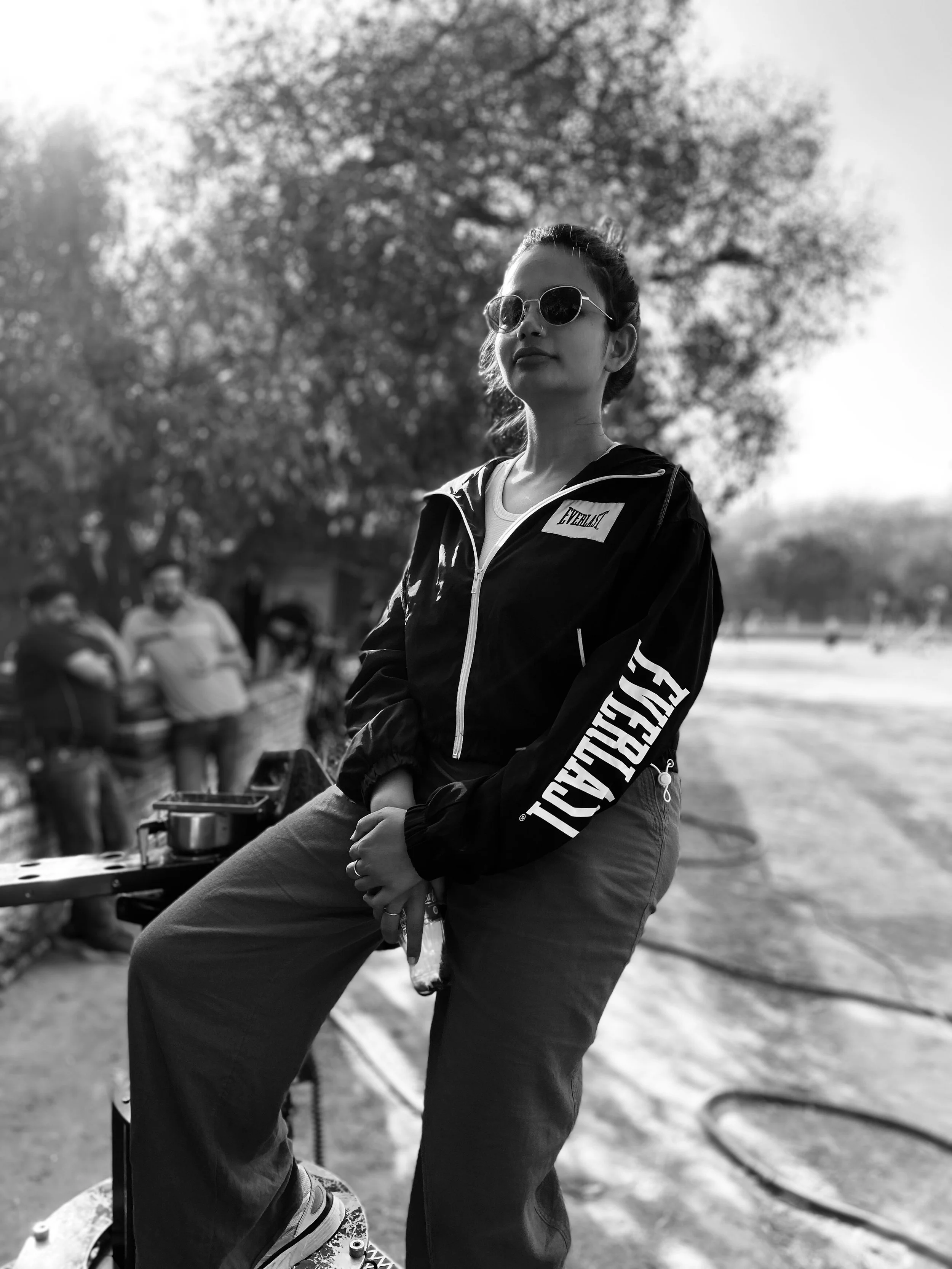 A woman with sunglasses standing outdoors near a tree, with a group of people in the background, in a black and white photo.