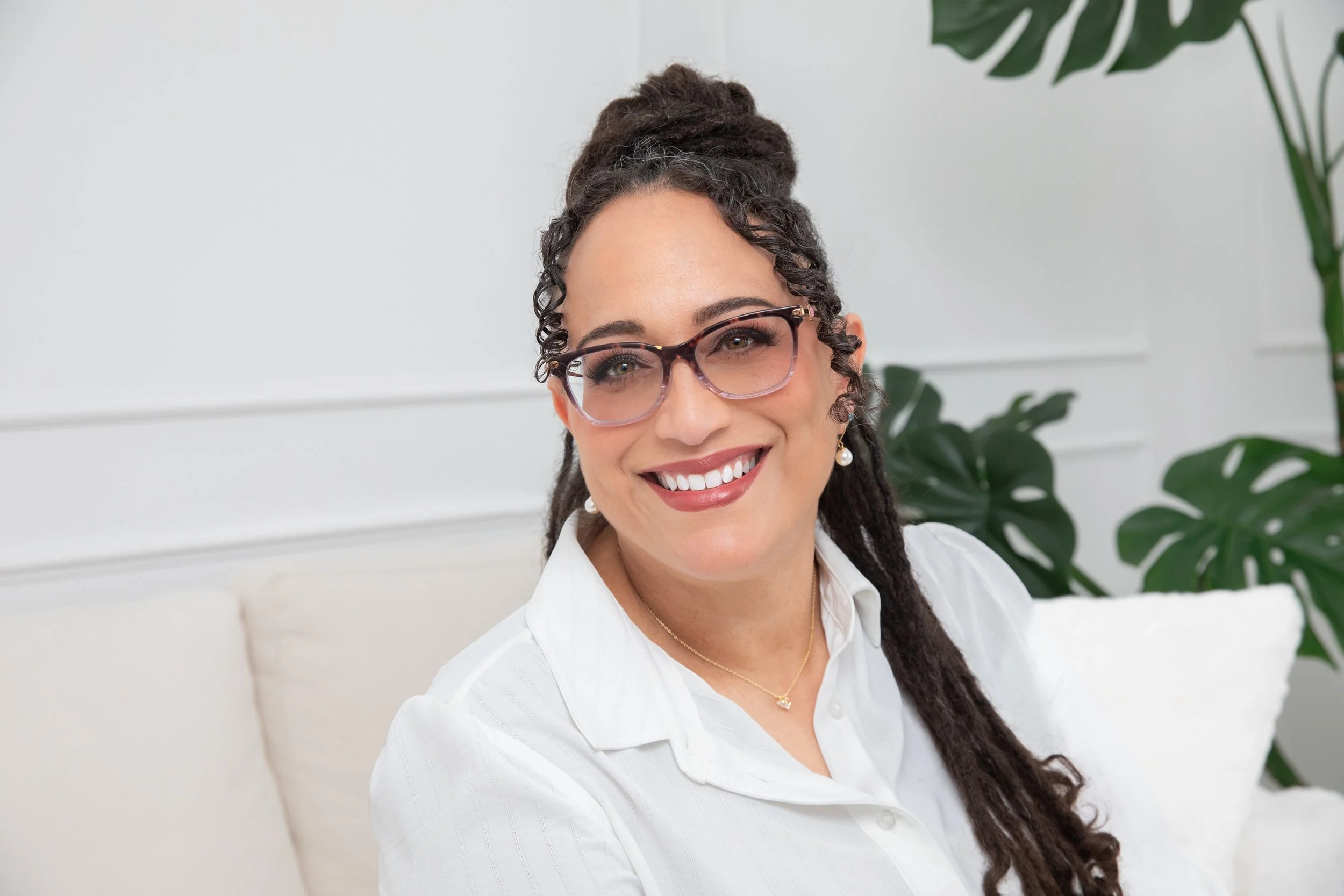 Nikki on the couch with hair half up, bold glasses and a brilliant warm and welcoming smile wearing a white collared blouse