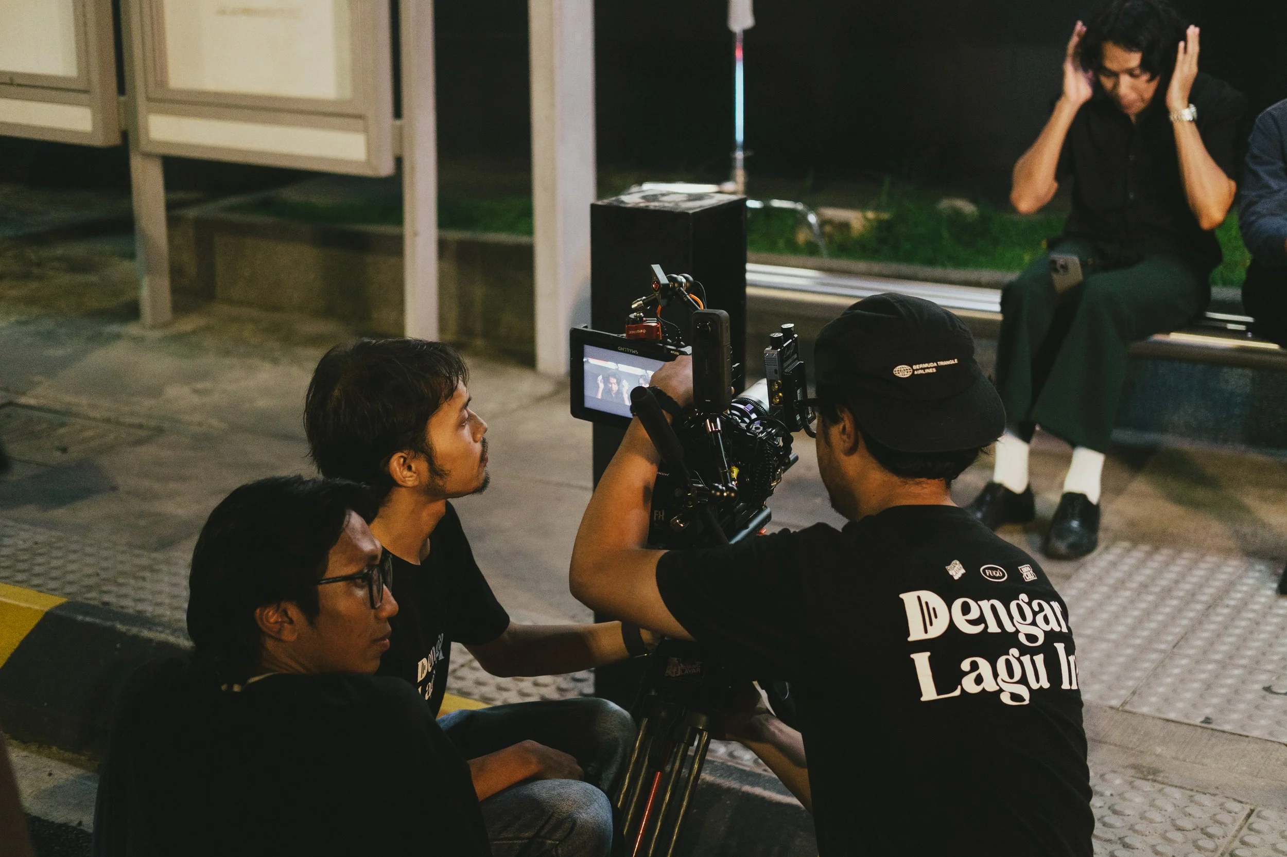 Filmmaker directing actors on a movie set at night, with a woman sitting on a bench in the background.