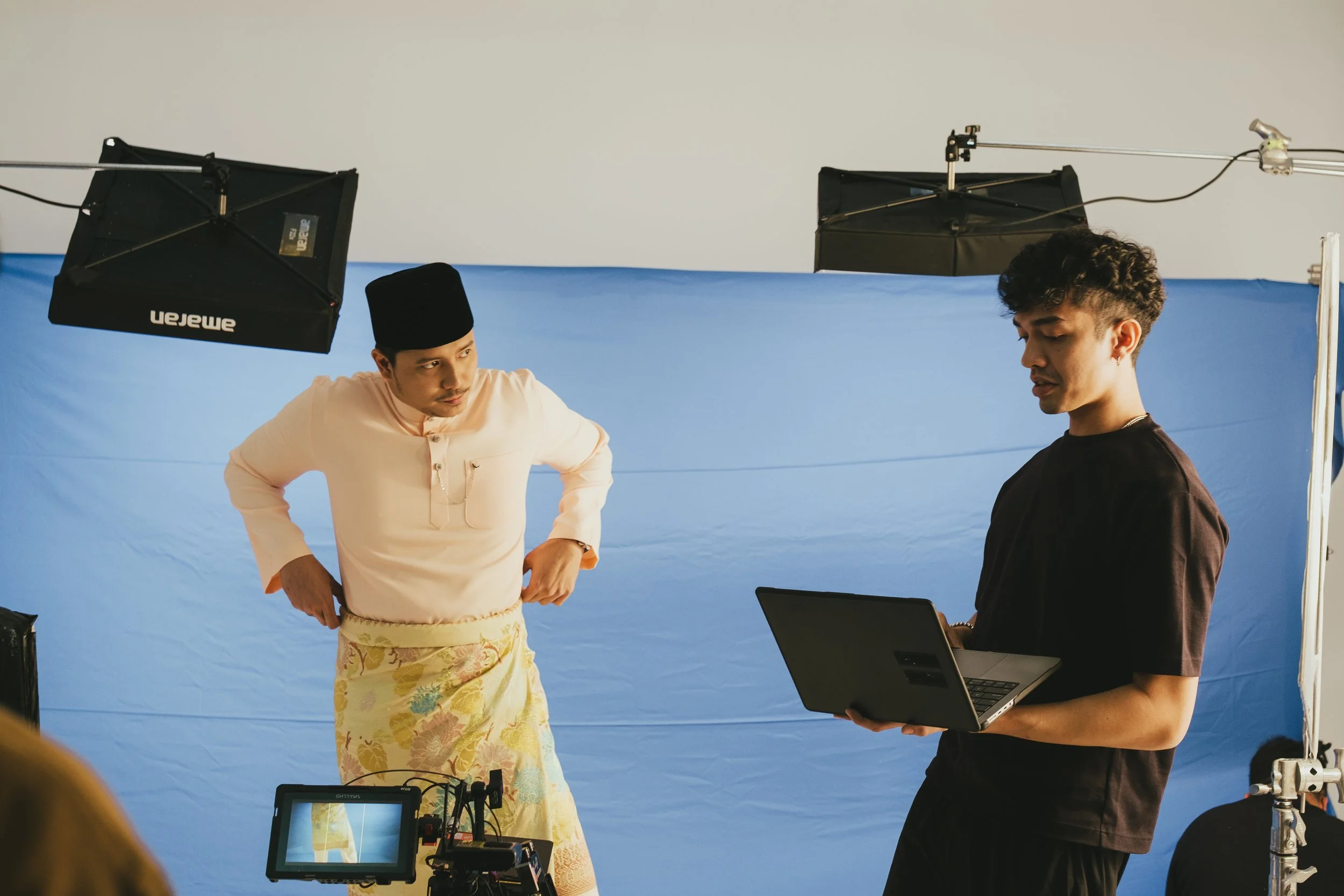A man wearing traditional Malay attire, including a songkok and a yellow sarong, stands in front of a blue screen during a filming session. Another man, dressed in black, holds a laptop and appears to be directing or reading from it. Studio lighting 