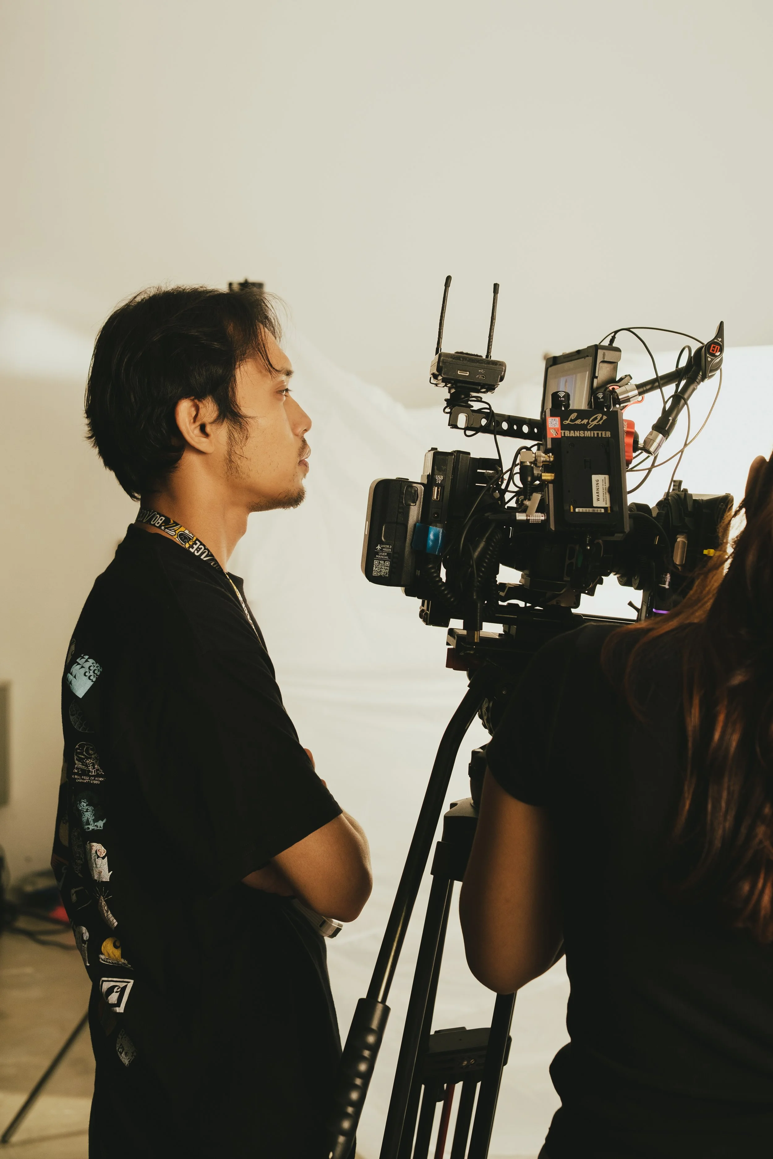 A man standing with arms crossed, looking at a professional video camera on a tripod, set up for filming.